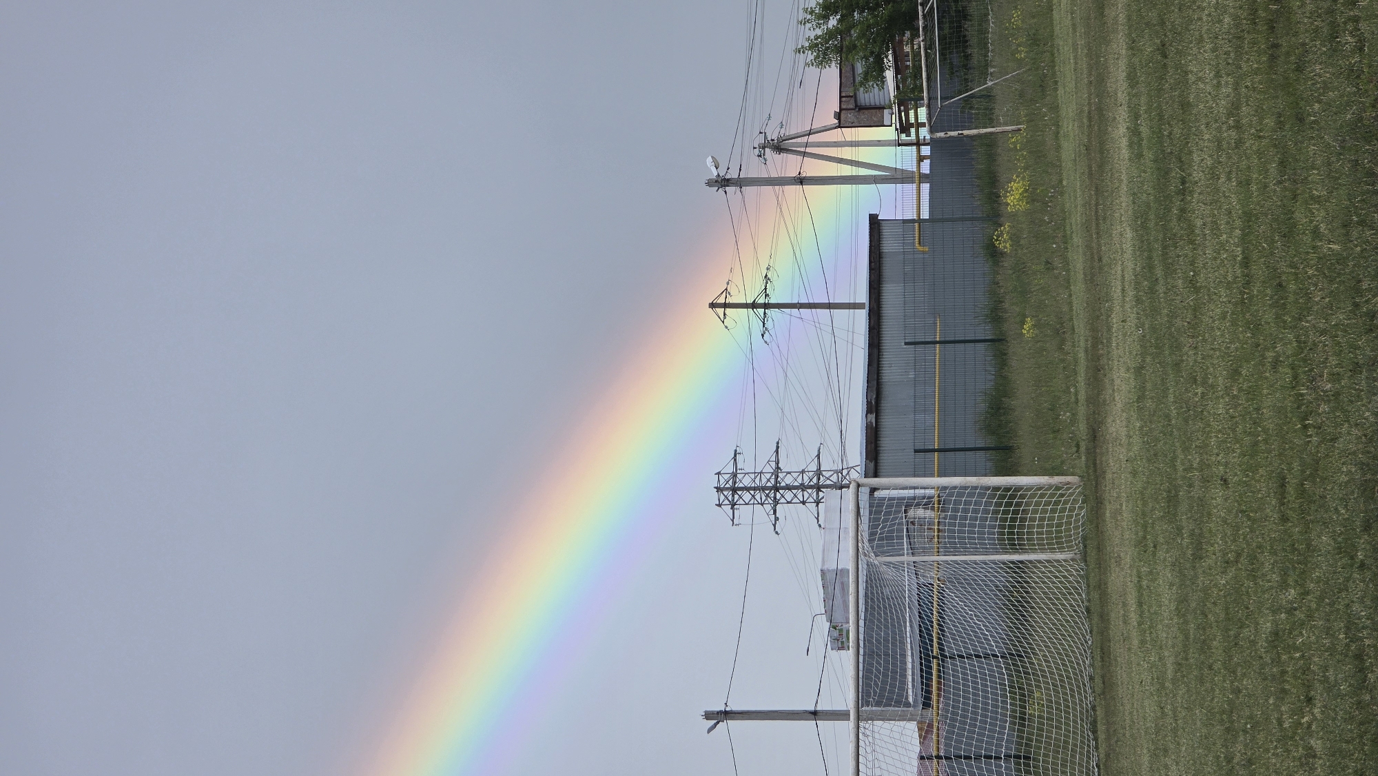 The image displays an outdoor scene featuring a vibrant rainbow stretching across a light blue-grey, overcast sky. The rainbow originates from the lower left and extends diagonally upwards towards the upper right, passing behind a series of utility poles with numerous power lines. In the foreground, on the right side of the image, there is a grassy field, likely a sports ground or football pitch, with a white-framed football (soccer) goal partially visible in the bottom right corner. Behind the utility structures and goal, industrial-style fences and parts of a building are visible, along with some distant green foliage. The presence of the rainbow indicates that it has recently rained or is currently raining, contributing to the diffused lighting. The image is oriented with the landscape rotated 90 degrees clockwise. No specific activity is depicted, and no visible text is present.