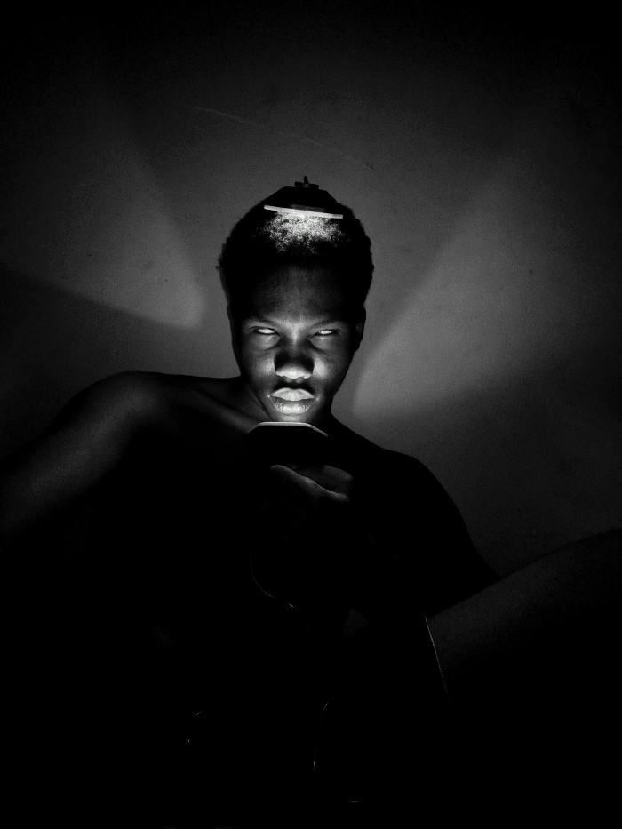 A monochrome, high-contrast image features a young Black person gazing intensely at the viewer. An object rests atop their head, illuminated with a bright light. Their eyes appear illuminated, and a hand holding a glowing phone or device casts light on their face and torso. The surroundings are shrouded in darkness, creating a dramatic, eerie effect. The composition emphasizes the subject's gaze and the play of light and shadow.
