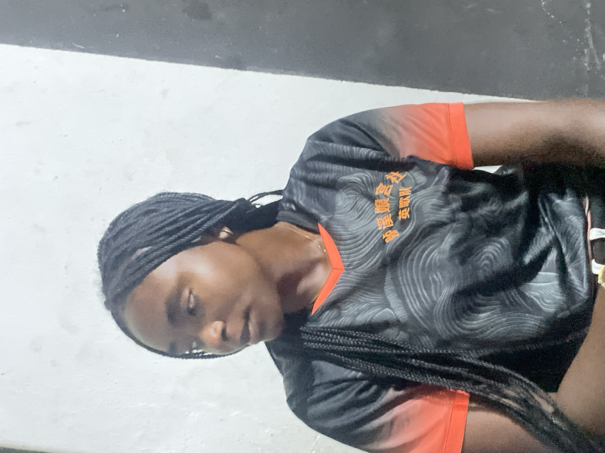 The image shows a young Black woman with braided hair. She is wearing a black and gray patterned shirt with orange sleeves and a V-neck. The shirt has orange text on the front, potentially in Chinese characters. She appears to be sitting or lying down on a light gray or white surface, possibly a floor. Part of a dark gray area is visible in the background. There is no visible information to determine if the image was taken in Jos, Nigeria.