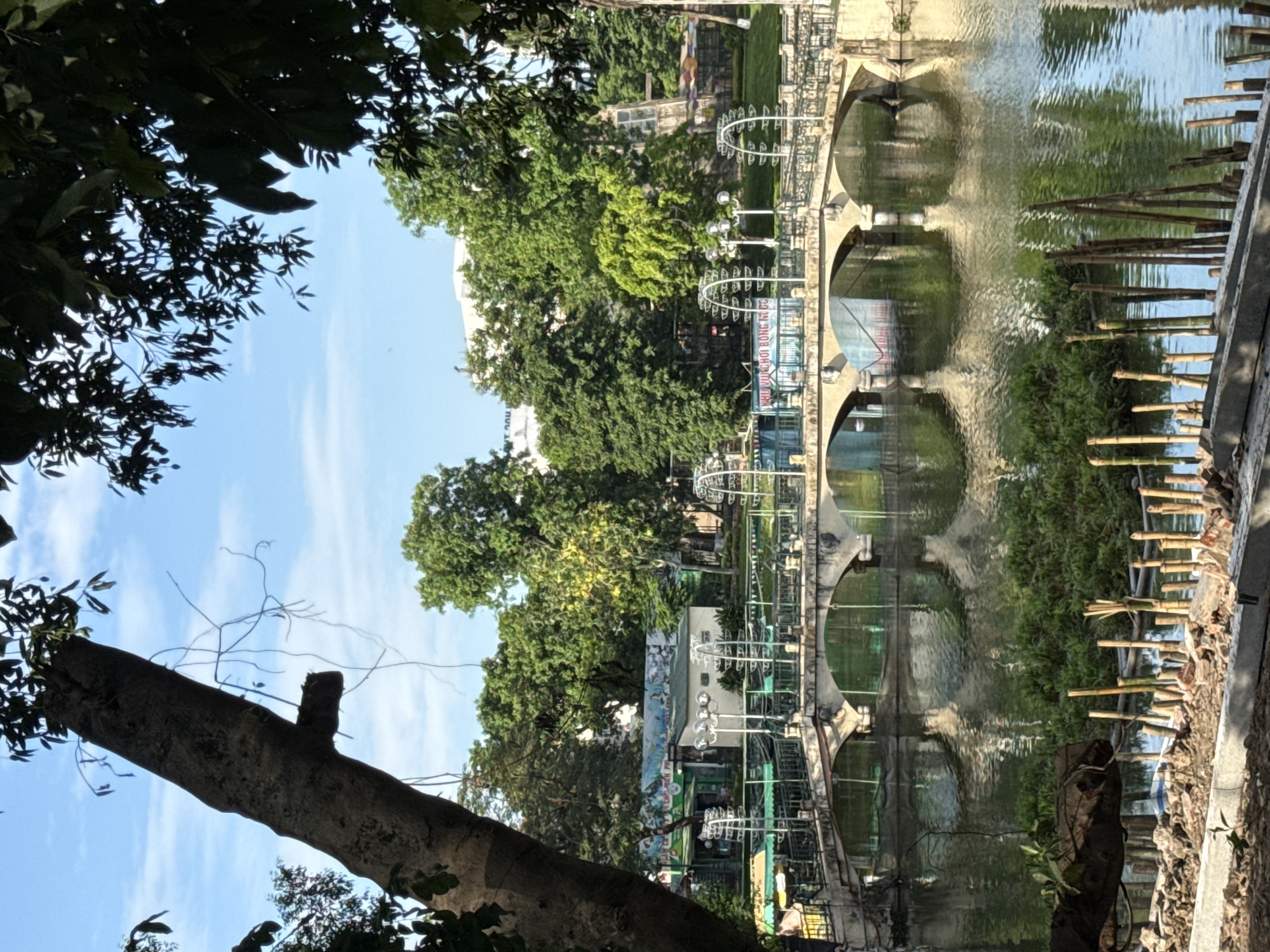 The image depicts a canal or pond in Hà Nội, Vietnam, with a bridge spanning the water. The bridge is constructed of concrete with archways. Adjacent to the water are bamboo structures and other elements, partially submerged. Trees and vegetation surround the canal, contributing to the park-like setting. The sky is visible through the trees, indicating a sunny day. Graffiti and art are noticeable on structures near the water. The image appears to be taken from a low vantage point, with a tree trunk and branches framing the view on the left. The reflection of the bridge and surrounding environment is visible in the water.