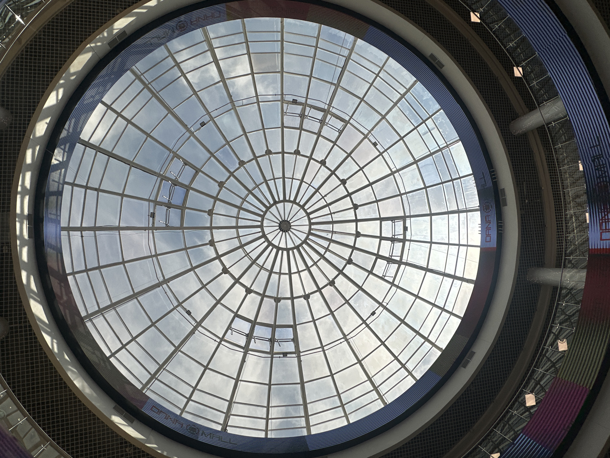The image shows a low-angle, wide shot of a large, circular, glass-domed ceiling, likely within a modern shopping mall or atrium. The dome is constructed from numerous geometric panes of glass, supported by a radial framework of metal beams that converge at a central point. Sunlight streams through the glass, illuminating the interior and casting shadows from the structural elements. In the lower portion of the image, sections of the mall's interior are visible, including tiered levels with railings and decorative facades featuring colored horizontal stripes and text that appears to be part of signage. A prominent sign reads "DANA MALL", indicating the location. The overall impression is one of modern architecture and open, light-filled space. This scene is located in the Dana Mall in Minsk, Belarus.