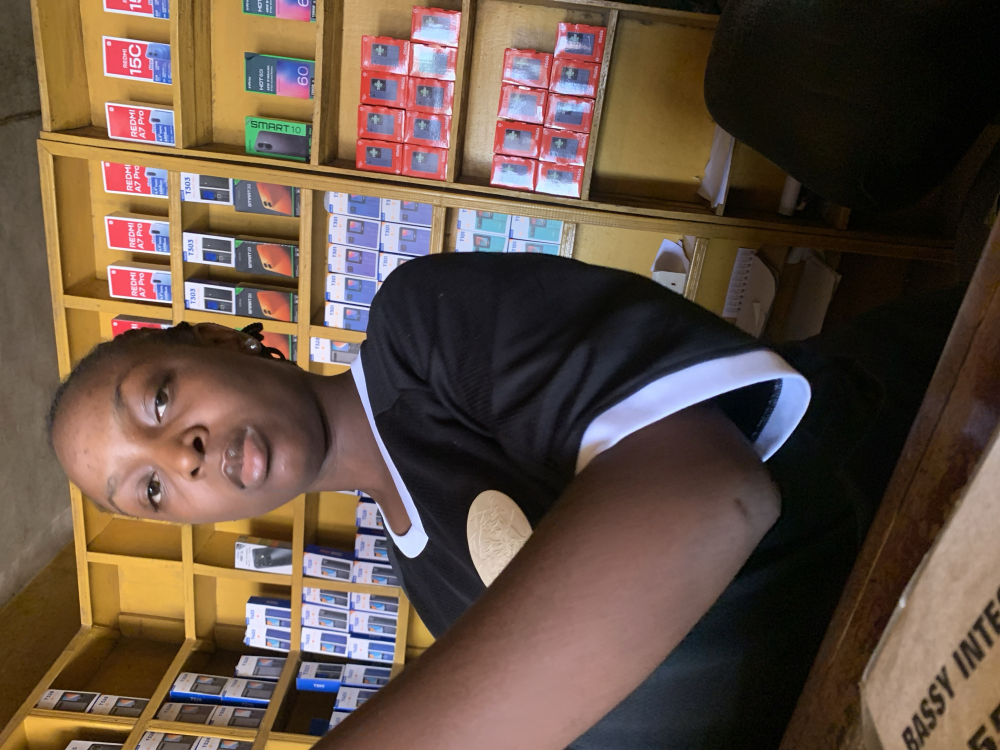 The image shows a person with dark skin wearing a black and white shirt, positioned in front of a shelf filled with boxes of mobile phones. The shelf appears to be made of yellow-painted wood and is stocked with various phone models, including "REDMI A7 Pro," "T303," "SMART 20," "T301," "REDMI 15C," and "SMART 10," among others. The boxes are arranged in rows and columns on the shelves. A small portion of a box labeled "BASSY INTE" is visible on the right. This scene seems to be taking place in a retail shop that specializes in selling mobile phones, possibly in Akwanga, Nigeria, given the context.