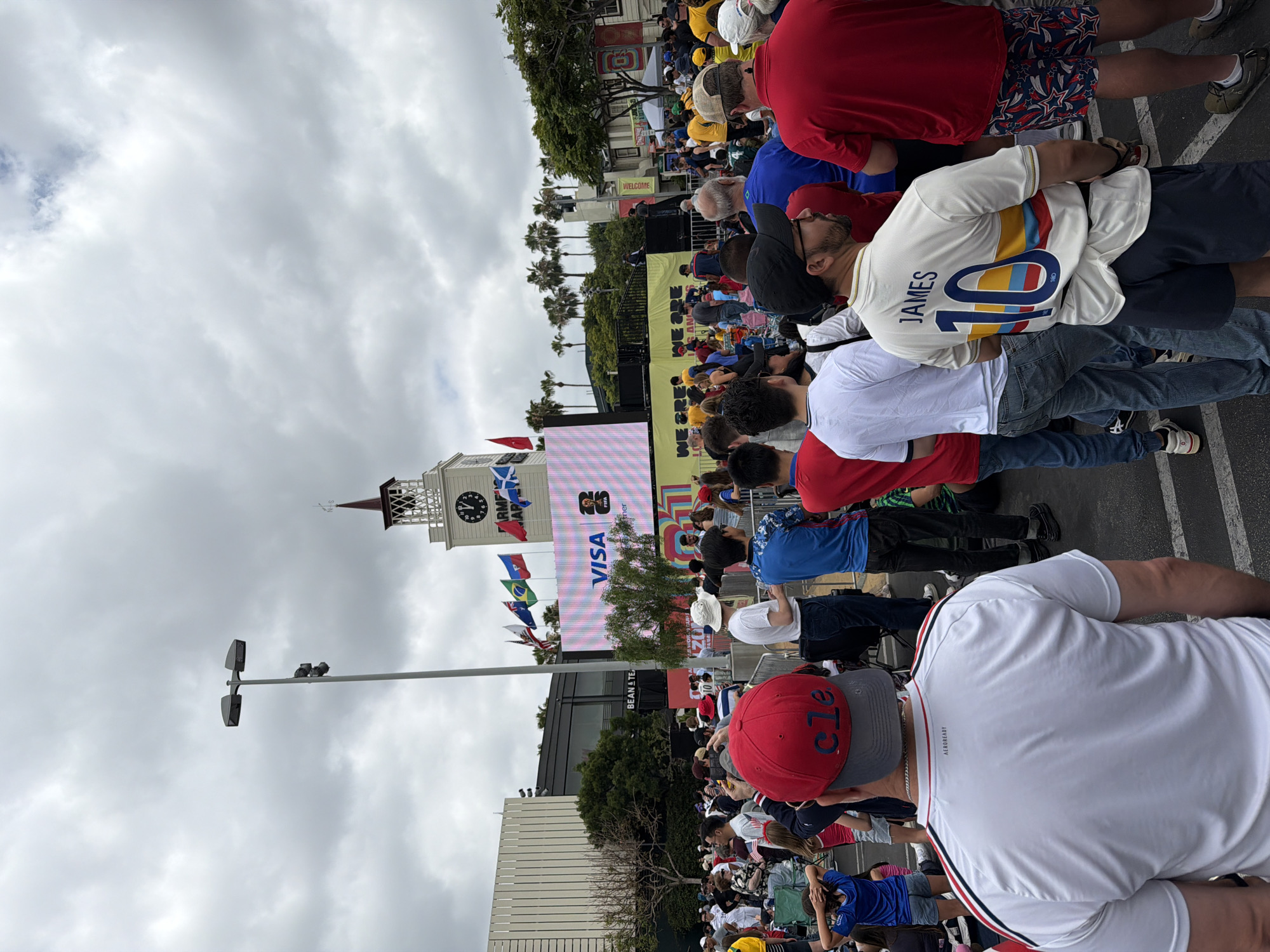 The image captures a large outdoor gathering of people, possibly for a sporting event or festival, with a clear view of a prominent clock tower and a large LED screen. The clock tower, located in the background, is white with a dark-colored spire and a weather vane. The LED screen displays the VISA logo and a FIFA World Cup graphic. Numerous flags, including the flags of Brazil, Scotland, and the United States, are visible, suggesting an international event. A crowd of people, many wearing casual attire and some with sports-themed clothing, are gathered in front of the screen and stage area. Palm trees line the background, and the sky is overcast. The scene appears to be taking place in an urban environment. The presence of the clock tower suggests a public square or a historic district within the city. The architecture and the type of gathering are consistent with events often held in the Pershing Square area of Los Angeles, particularly during major international sporting events.