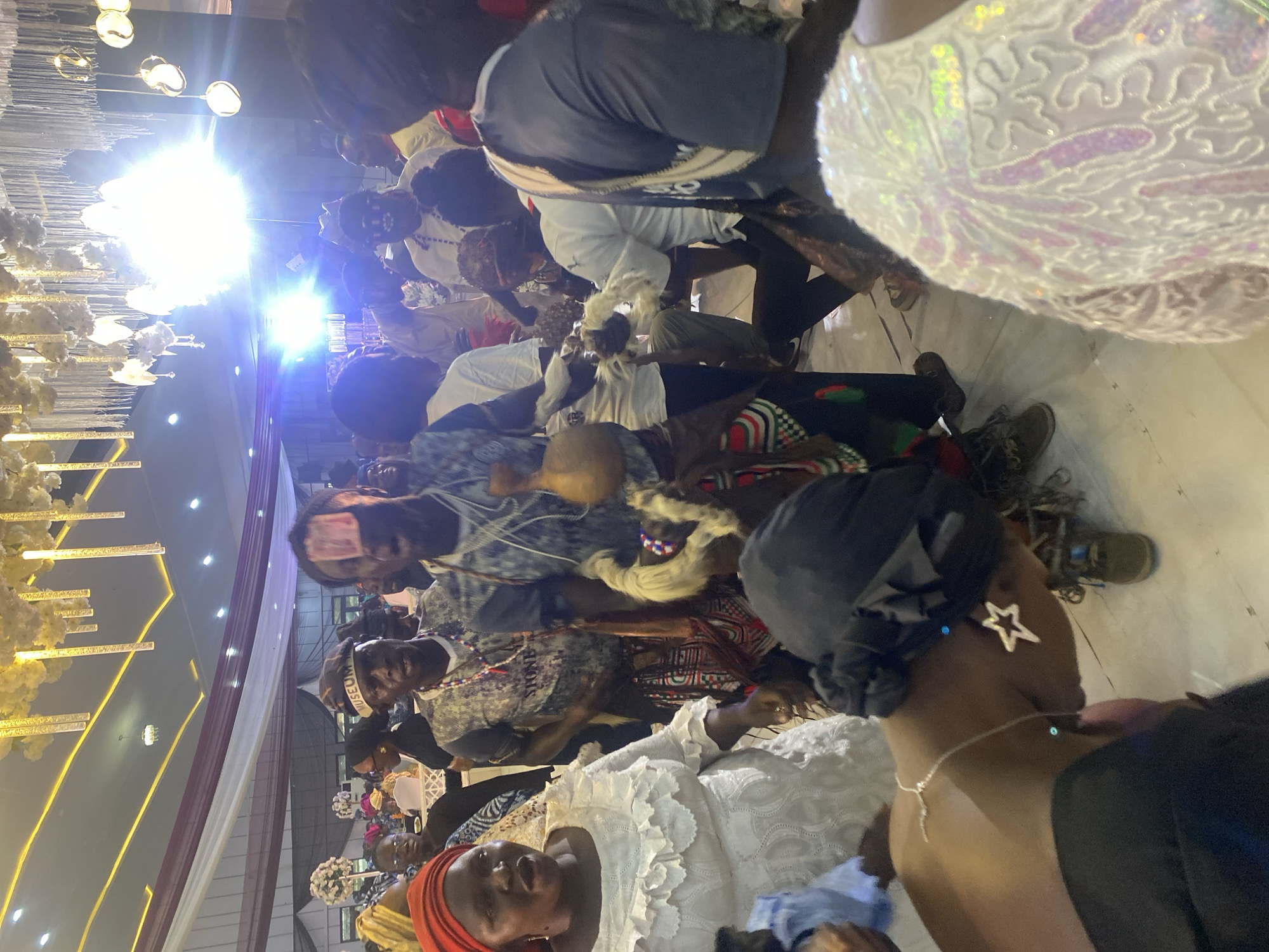 The image shows a gathering of people in an indoor setting, likely a hall or event space. There appear to be several individuals dressed in traditional attire, suggesting a cultural event or celebration. The location is possibly in the city of Jos, Nigeria, but this cannot be confirmed with certainty from the image alone.
People:
- Multiple individuals of varying ages and genders are present.
- Some are dressed in traditional Nigerian clothing, including head wraps, patterned fabrics, and ornate jewelry.
- One individual has what looks like a bank note placed on their forehead.
- One person is wearing a black head covering and has a star shaped earring.
- Many are wearing jewelry, including beaded necklaces.
Objects:
- A chandelier is visible in the background.
- Decorative elements such as flowers and draping fabrics are present.
- At least one person holds a carved wooden object.
Actions/Interactions:
- People are seated and appear to be interacting with one another.
- Some individuals are gesturing with their hands.
Environment:
- The setting is a well-lit interior space with decorated columns.
- The floor appears to be tiled.
- The room is crowded with people.
