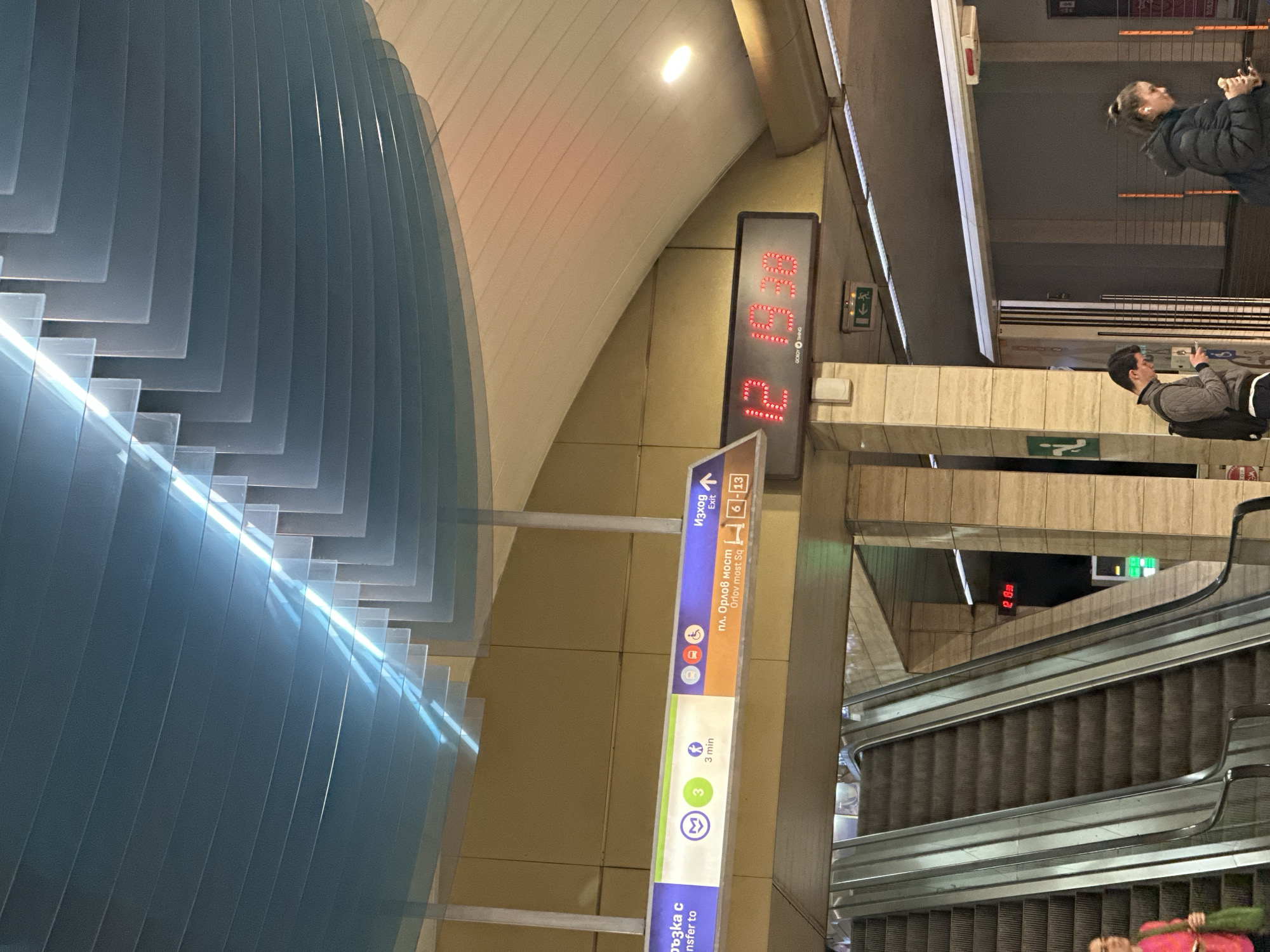 The image is rotated 90 degrees counter-clockwise, showing an interior view of a modern metro station, likely an underground concourse or transfer area, during the evening.
On the left, a prominent architectural feature is visible, composed of multiple layered, translucent panels in shades of blue and grey, forming a curved wall or ceiling element. Streaks of light reflect across these surfaces.
In the center of the frame, a tall, vertical directional sign provides information in both Cyrillic and English. The top section indicates "Изход" (Exit) with an upward arrow and numbers "6-13". Below this, it directs to "пл. Орлов мост" (Orlov most Sq), a well-known location in Sofia, Bulgaria, accompanied by a walking icon suggesting a 3-minute journey. The bottom section shows "ръзка с" (Transfer to) metro line 3, represented by a green circular 'M' icon.
Adjacent to the sign, a large digital display shows "12 19:30", indicating the time as 7:30 PM (19:30) and potentially a platform or track number "12". The word "COMING" is also visible on this display.
To the right, a set of escalators descends into the lower levels of the station. Several people are present; a person in a dark jacket is near the top of the escalators, looking at a phone. Further down, another person in a grey jacket with a backpack is also focused on a phone. A third person, partially visible at the bottom of the escalators, is wearing pink attire.
The station features clean, tiled walls and artificial lighting, contributing to the impression of a functional urban transit environment.