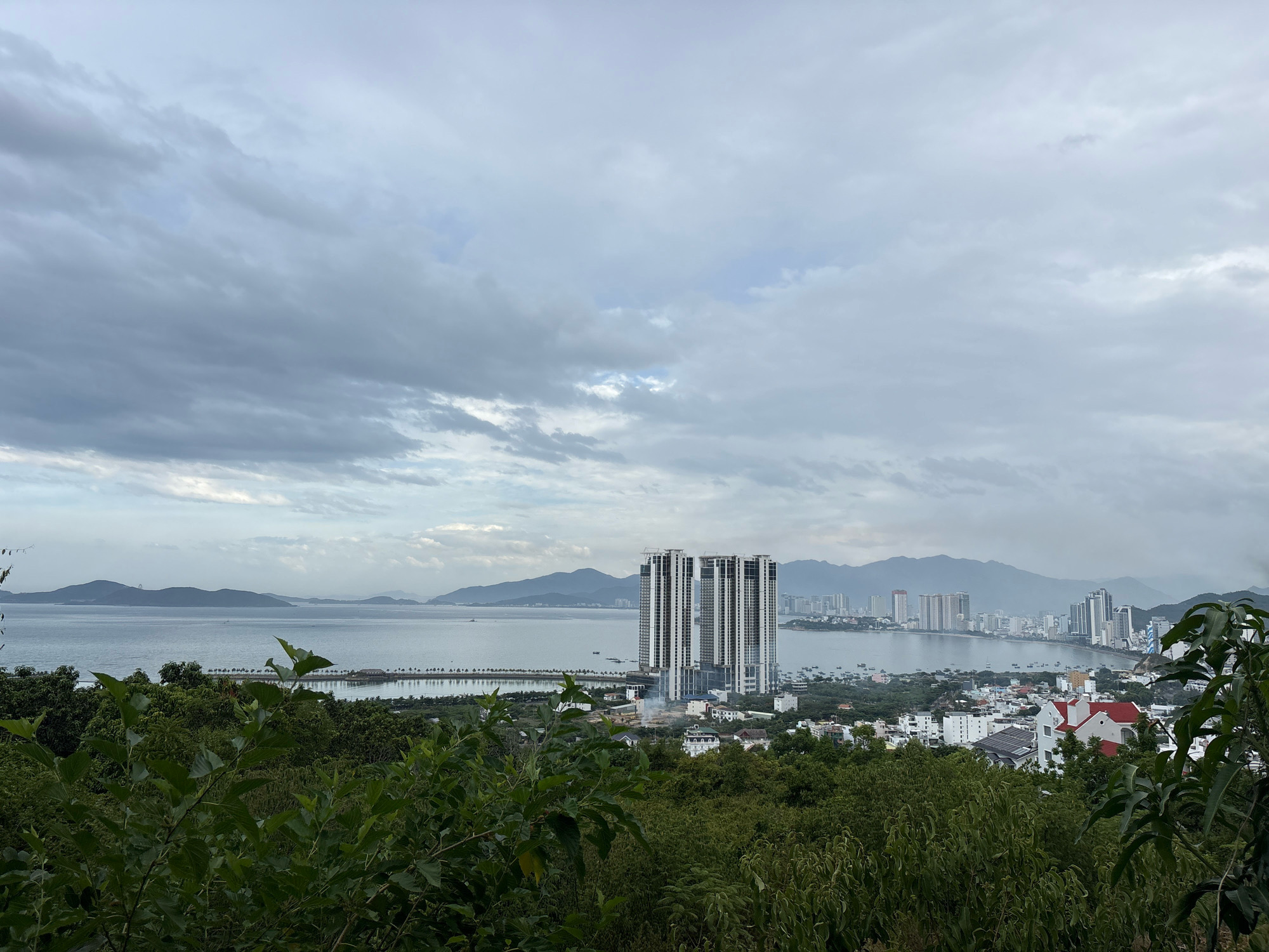 The image depicts a panoramic view of a coastal city, likely Nha Trang, Vietnam, under a cloudy sky. In the foreground, lush green foliage partially obscures the view. The mid-ground showcases a body of water, possibly a bay or ocean, with a long pier or breakwater extending into it, lined with palm trees. Several boats are visible on the water. Dominating the cityscape are clusters of modern high-rise buildings, including a prominent set of three interconnected skyscrapers with a distinctive architectural design. Further inland, lower-rise buildings, some with white facades and red roofs, are scattered among the trees. In the background, a range of hazy, mountainous terrain stretches across the horizon. The overall impression is of a developing urban coastal environment.