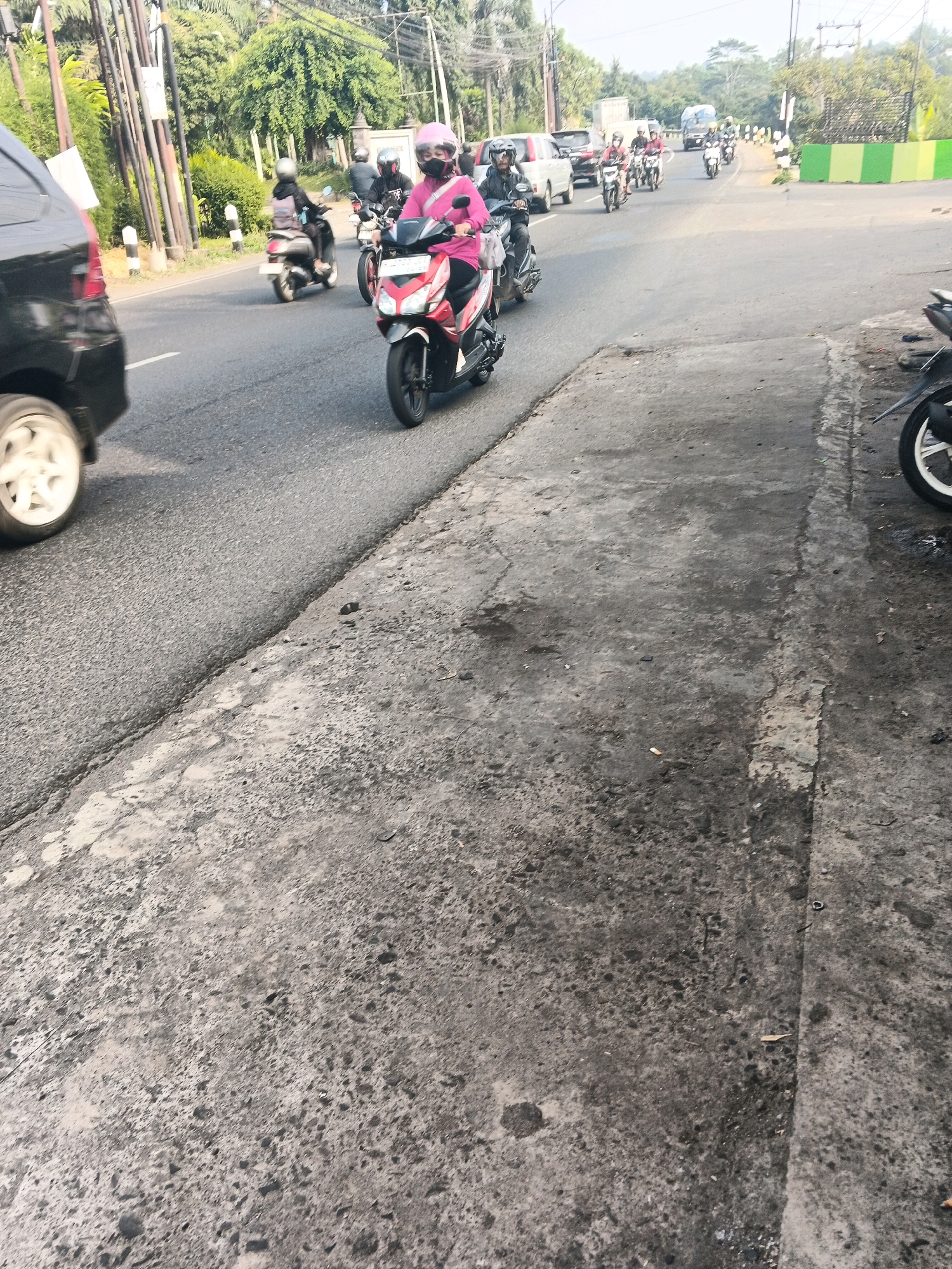 A group of motorcyclists are traveling on a road in Ungaran, Indonesia. The scene depicts a typical street in Ungaran with a paved road, sidewalks, trees, and electrical poles lining the sides. In the foreground, a black car is partially visible on the left, with its wheel on the road. Several people on motorcycles are in motion, with one rider in a pink jacket and helmet prominently featured. The motorcyclists appear to be navigating traffic on a sunny day, with the background showing more vehicles and lush green vegetation. The overall impression is of a busy urban environment with a significant presence of motorcycle transportation.