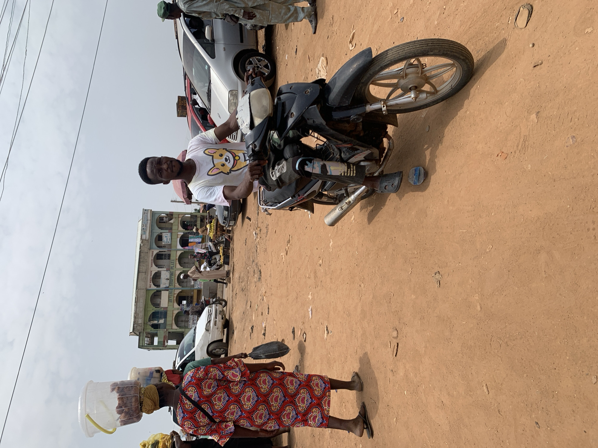 The image captures a street scene in Akwanga, Nigeria. A man is seated on a black motorcycle, wearing a white t-shirt with a cartoon dog print. In the foreground, a woman is carrying a container on her head, walking leftward. She is wearing a red patterned dress. The ground is dirt, and there are cars and a building in the background. Overhead wires are visible in the upper left corner. Other people are present in the background.