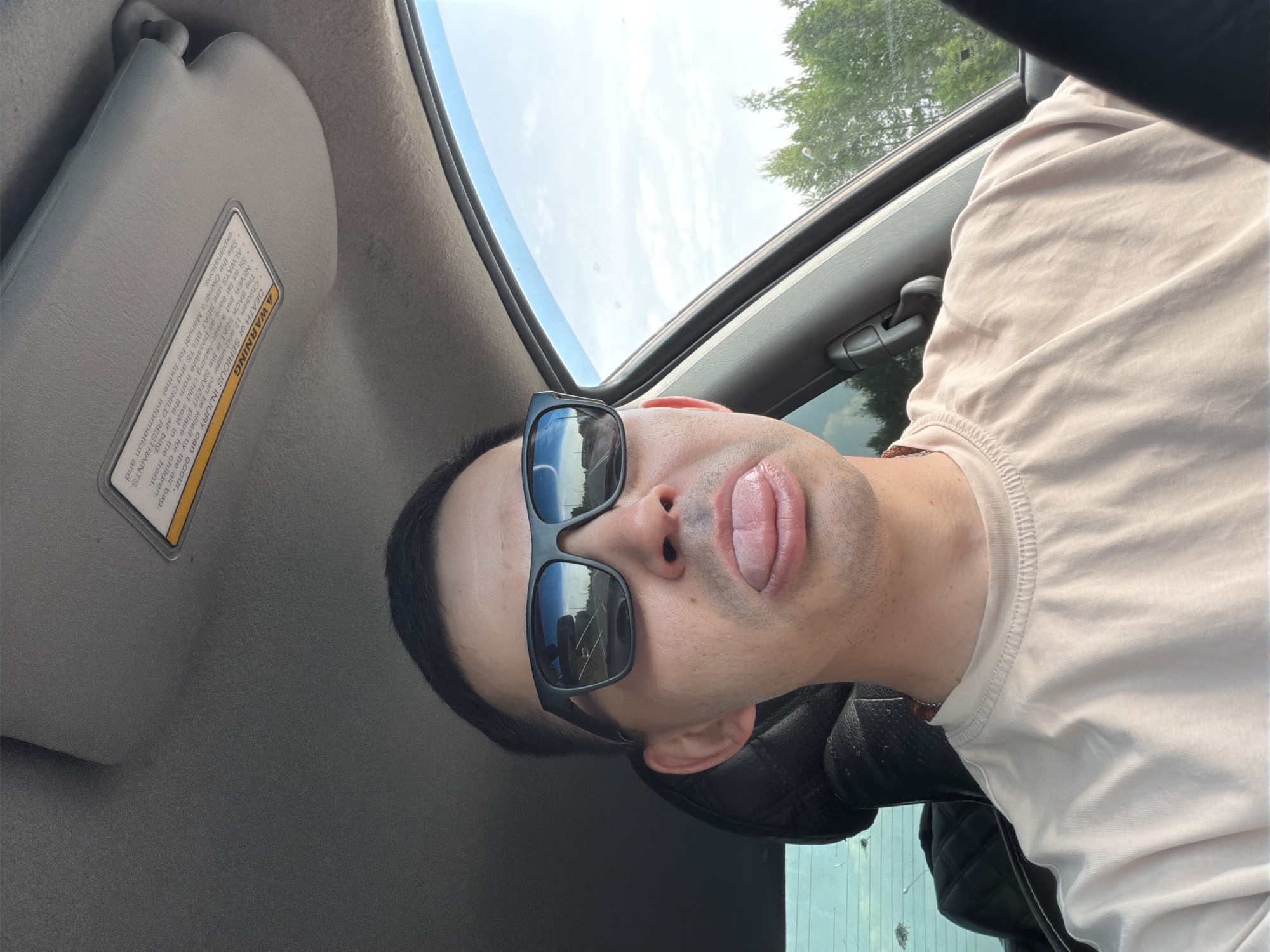 The image is a selfie taken from inside a car. A person wearing sunglasses and a light-colored shirt is sticking their tongue out. The interior of the car is visible, including the sun visor with a warning label, the sunroof, and a black quilted leather seat. The sky is visible through the sunroof, with some trees in the background.