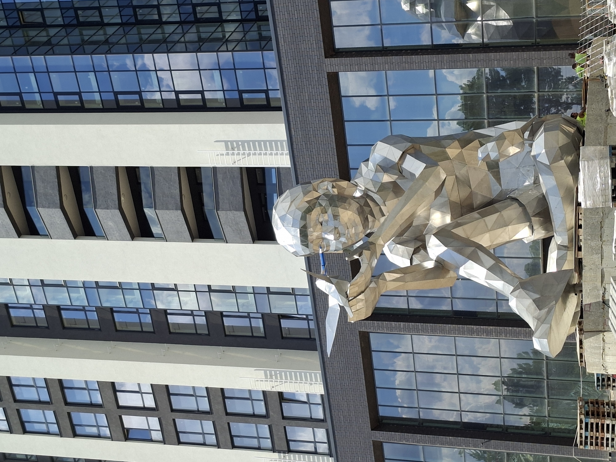 A large, polygonal, metallic sculpture of a child holding a bird is positioned in front of a modern building with a facade of dark brick and large glass windows reflecting the sky. The sculpture is made of many geometric facets, giving it a faceted, crystal-like appearance. The child is depicted sitting with its legs crossed, and its head is tilted slightly upwards as it gazes at the bird in its hands. In the background, the building features alternating bands of light-colored and dark gray material, with prominent vertical white elements. The glass windows on the building reflect the blue sky with scattered clouds and trees. The sculpture is situated on a concrete base, surrounded by construction materials such as wooden pallets and plastic wrap. Several construction workers in yellow vests are visible in the lower right portion of the image. This sculpture is located in the city of Chișinău, Moldova, specifically at the intersection of Ștefan cel Mare și Sfânt Boulevard and Armenească Street, in front of the new office building "Efcom Tower".