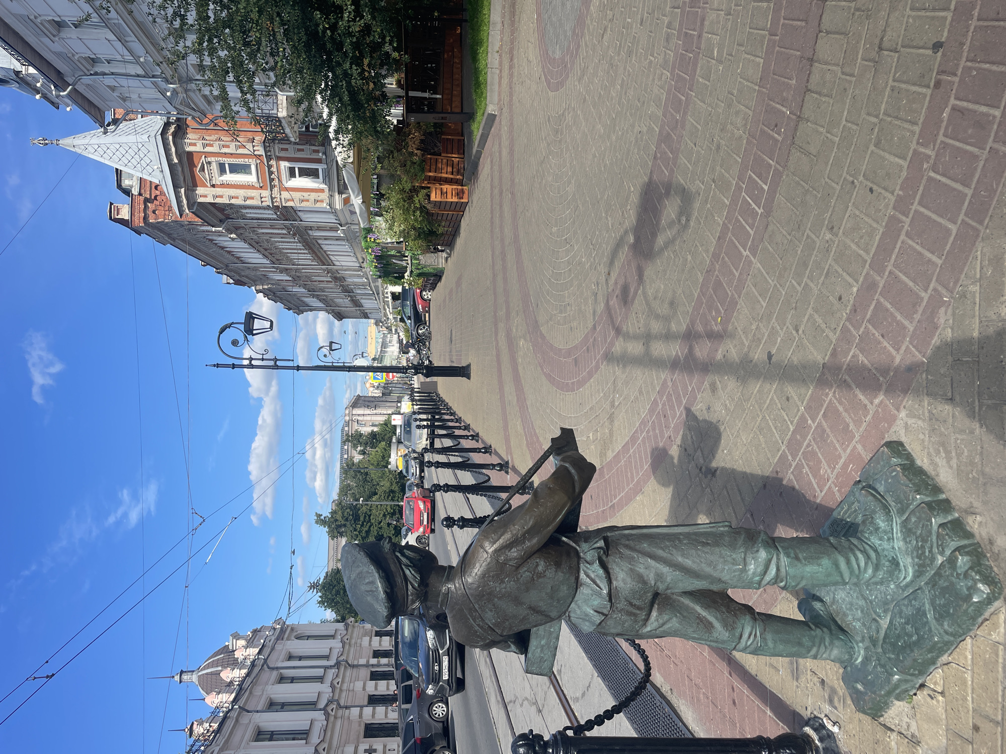 The image depicts a street scene in Nizhny Novgorod, Russia. In the foreground, a bronze statue of a boy in uniform, possibly a soldier or a messenger, stands on a stone pedestal. He is wearing a cap and holding a box. The statue is positioned on a paved sidewalk with a repeating pattern of brickwork. To the left of the statue, there is a black metal fence with chains, and beyond that, tram tracks and several cars on the road.
The street is lined with historical buildings with ornate architecture, typical of the city center. Some buildings have multi-story facades with various window designs and rooflines, including a prominent building with a distinctive spire. The street is well-lit by streetlights. There are awnings extending from some of the buildings, suggesting commercial establishments like cafes or shops. Green trees and bushes are visible along the sidewalk and in front of some buildings. The sky is blue with scattered white clouds, indicating a clear, sunny day. The overall impression is a charming and historic urban environment.