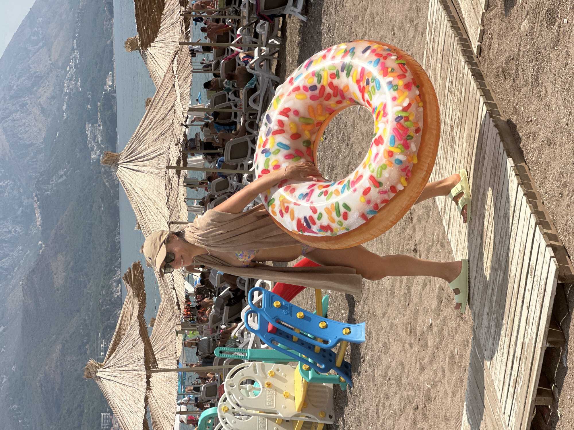 A woman walks on a wooden path on a sandy beach, carrying a large inflatable donut-shaped ring. She is wearing a light-colored towel draped over her shoulders, a bikini, a baseball cap, and sunglasses. In the background, beach umbrellas, lounge chairs, and people are visible. Beyond the beach, the sea is calm, and a mountain range with buildings on its slopes forms the backdrop, suggesting the location is Boreti, Montenegro. A small children's play structure with slides is also present on the left side of the image. The scene depicts a sunny day at a crowded beach resort.