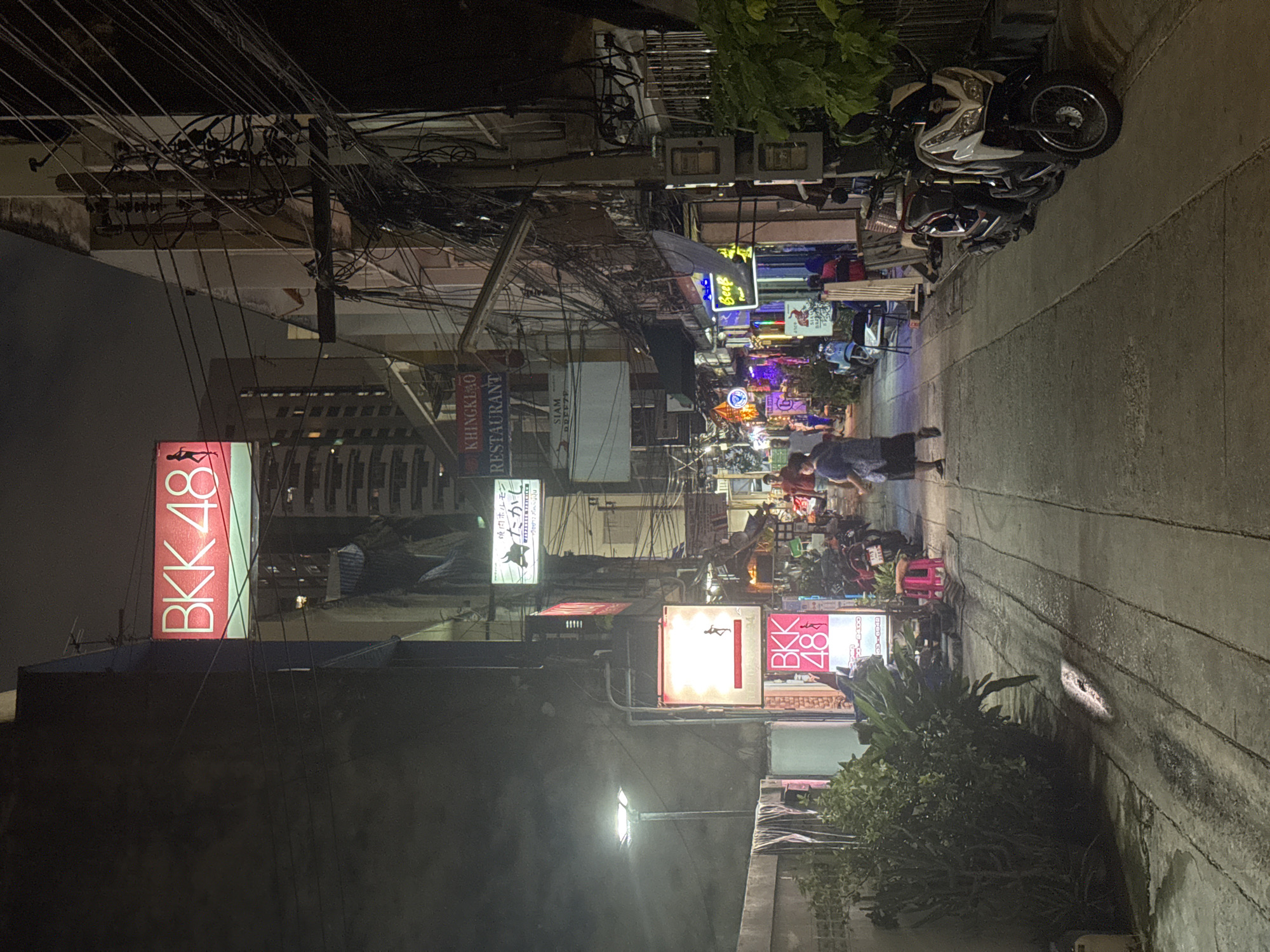 This is a vertical, nighttime photograph of a bustling street scene in Bangkok, Thailand. The scene is packed with numerous signs, many of which are illuminated, indicating a commercial area with restaurants and shops. Electrical wires are densely strung overhead, a common sight in many parts of Bangkok.
In the foreground, a large, red and white neon sign reads "BKK 48". Further down the street, other signs with Thai and English text are visible, including "KHINGKLAO RESTAURANT", "SIAM", and "Beef". There are also Japanese characters on some signs, such as "たかし".
Several motorcycles are parked along the sidewalk, and a few people are walking or standing. One person in a dark shirt and shorts is walking away from the camera on the right side of the street. Another person, wearing a red shirt, is visible in the background. The street appears to be paved, and there are planters with lush green foliage lining parts of the sidewalk. The overall impression is a vibrant, energetic street at night, characteristic of a lively urban environment in Bangkok. The lighting suggests it is well after dark, and the atmosphere seems active and engaging.