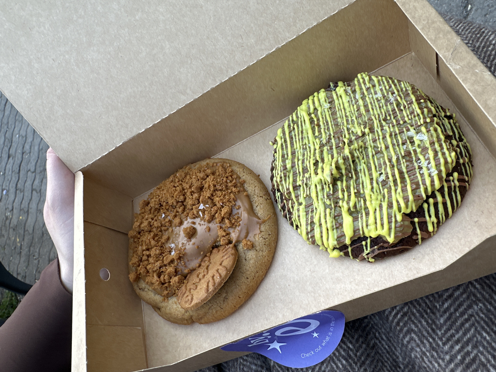 The image shows a cardboard box containing two large cookies. The cookie on the left has a light brown base, topped with a smooth caramel-like spread, crushed cookie crumbs, and a whole Biscoff cookie. The cookie on the right is chocolate-based, drizzled with a bright yellow glaze and sprinkled with salt crystals. A hand holds the box open. A blue and white sticker is visible on the lower edge of the box. The location is Sofia, Bulgaria.