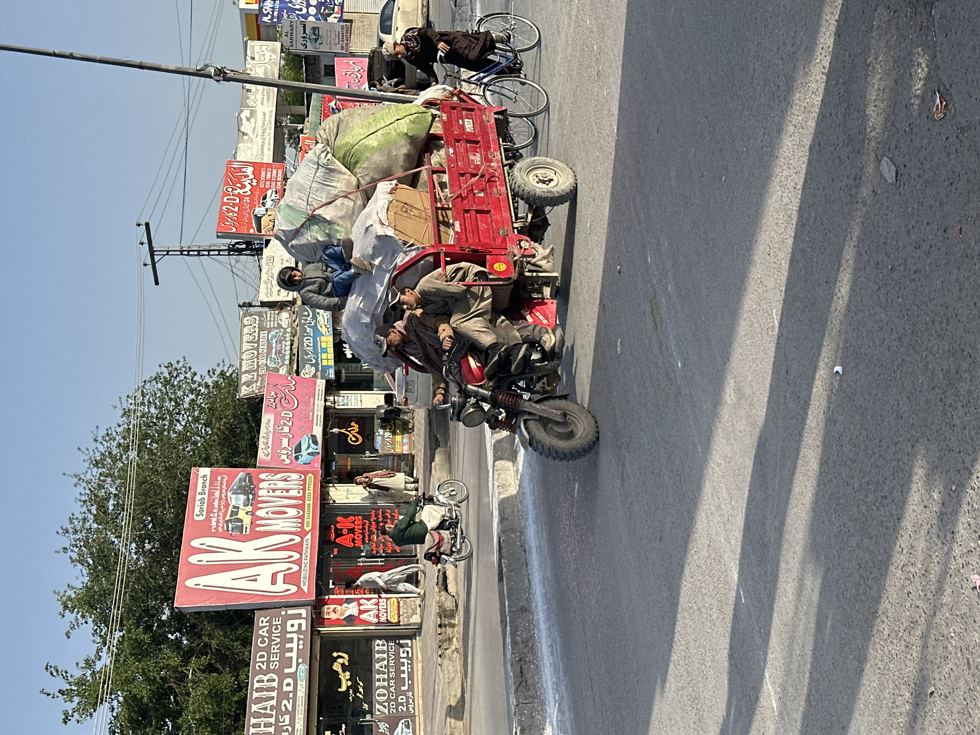 The image captures a vibrant street scene in what appears to be a commercial district, likely in Quetta City Tehsil, Pakistan. The central focus is a red three-wheeled cargo vehicle, seemingly a modified motorcycle rickshaw or a motorcycle with an attached trailer, heavily laden with large, irregularly shaped bundles or sacks, some appearing to be filled with soft materials. A cardboard box is also visible among the cargo. Two men are on the vehicle; one is positioned as if driving the attached motorcycle, while another sits amidst the cargo in the back.
In the background, a person in dark clothing is pushing a bicycle. The street is lined with numerous colorful billboards and signs, indicating a busy commercial area. A utility pole with power lines runs across the upper part of the frame, and lush green trees are visible further back. The bright, clear sky and distinct shadows cast on the road suggest it is daytime, likely mid-morning or late afternoon, under sunny weather conditions.
The visible text on the signs, predominantly in Urdu, provides strong location cues. Several signs advertise "AK MOVERS" with the tagline "MOBILIZING PATHWAYS" and offer "All Pakistan service in more than 50 cities," mentioning a "Sariab Branch." Other prominent businesses include "ZOHAIB 2D CAR SERVICE" and "AL SARWARY." Phone numbers are also displayed on various advertisements. The overall setting depicts everyday activity in a bustling South Asian urban environment.