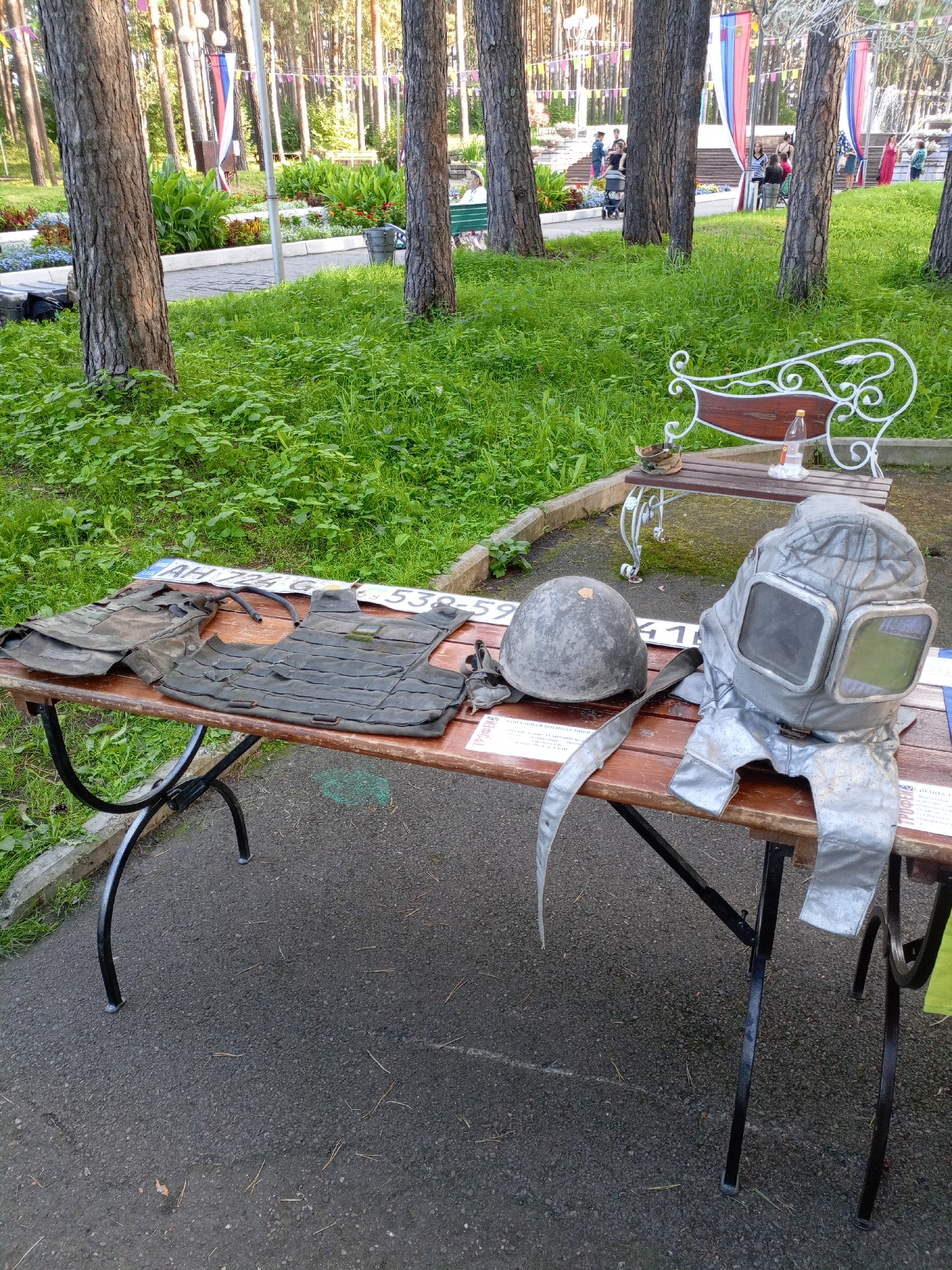 In Zheleznogorsk, Russia, a wooden table displays a collection of military and protective gear in a park-like setting. On the left, two dark green tactical vests are laid out, one appearing partially disassembled. Next to them are what seem to be license plates with Cyrillic lettering and numbers. To the right of the vests, a metallic grey helmet rests on the table, and to its right, a silver-colored, futuristic-looking helmet with large, clear visors is prominently displayed. A pair of black gloves is also visible on the table, near the helmet. The table itself has black metal legs and is positioned outdoors on an asphalt surface, with a grassy area and trees in the background. In the further background, people can be seen in a park area with decorative flags and foliage. A white ornate bench with a wooden seat is situated behind the table, with a plastic bottle and a small container on it.