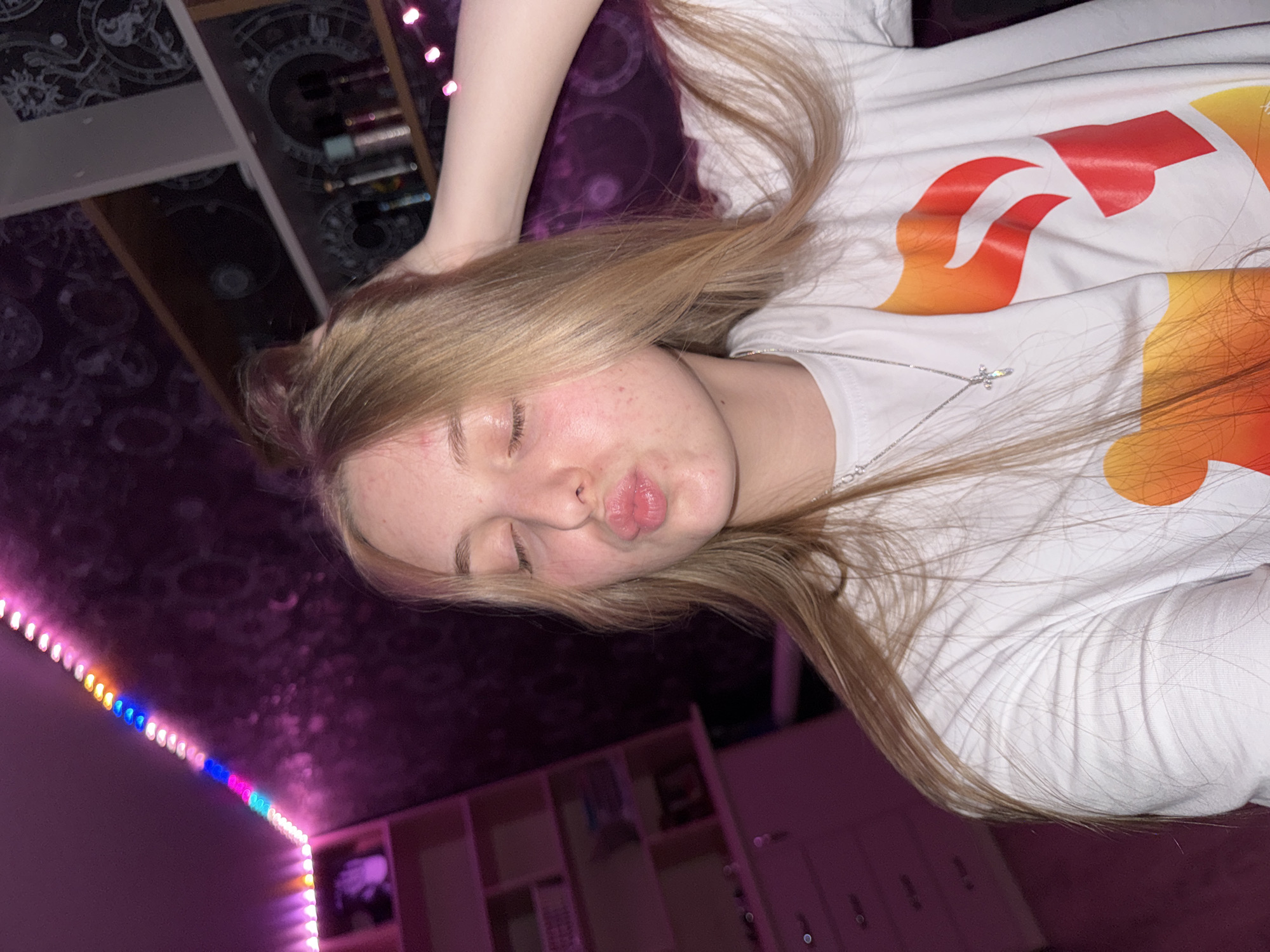 A young Caucasian female with blonde hair is shown from the chest up, looking into the camera with her eyes closed and puckering her lips. She is wearing a white t-shirt with an orange and red graphic design and a silver cross necklace. Her hand is placed in her hair. The background is a bedroom with pink LED strip lights, a shelf with bottles and decorative items, and a purple patterned wallpaper. A pink dresser is visible on the left side of the image.