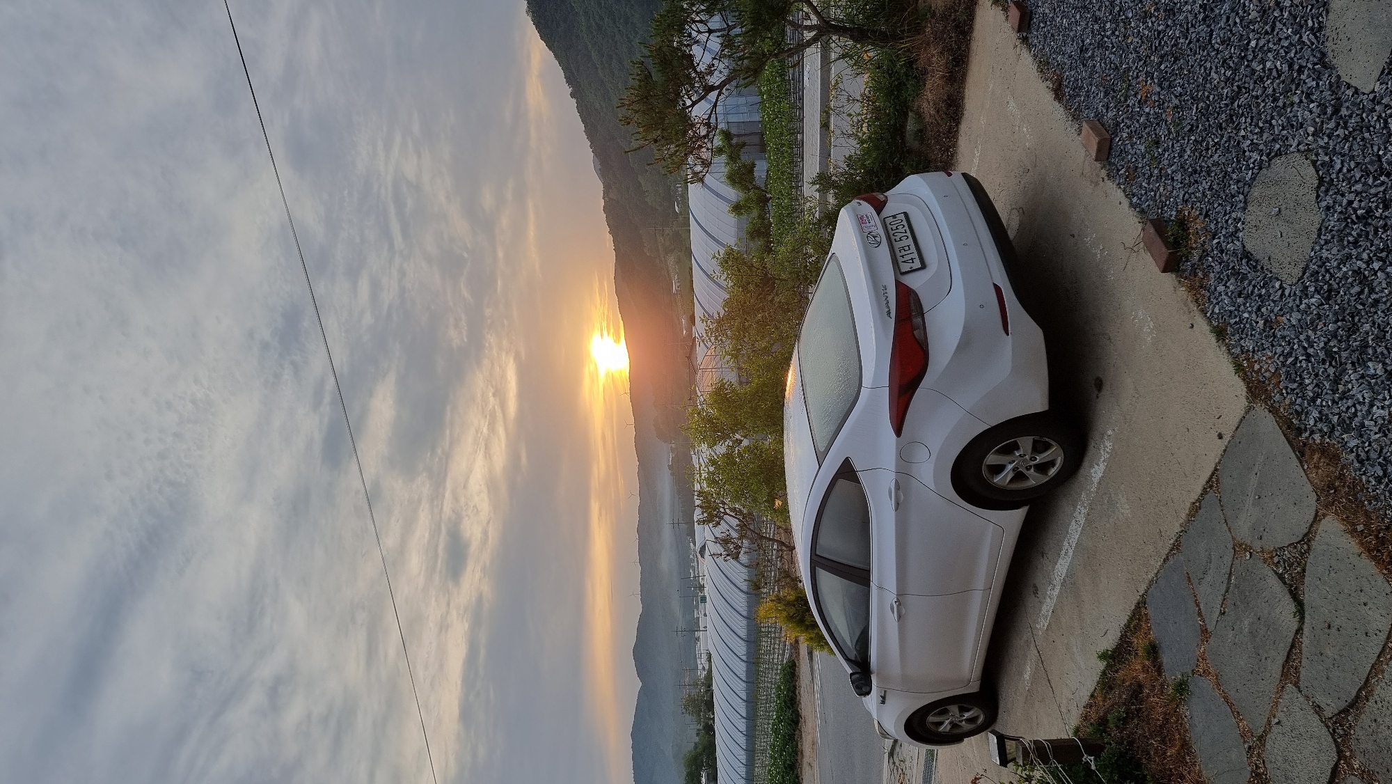 A white Hyundai Elantra sedan is parked on a concrete road next to a pebble-covered area. The car is facing towards the right of the image. Behind the car are greenhouses and trees. In the background is a mountain range with the sun setting. There is a power line in the sky. The car's license plate reads "47라 5250". The image was taken in Dunnae-myeon, South Korea.