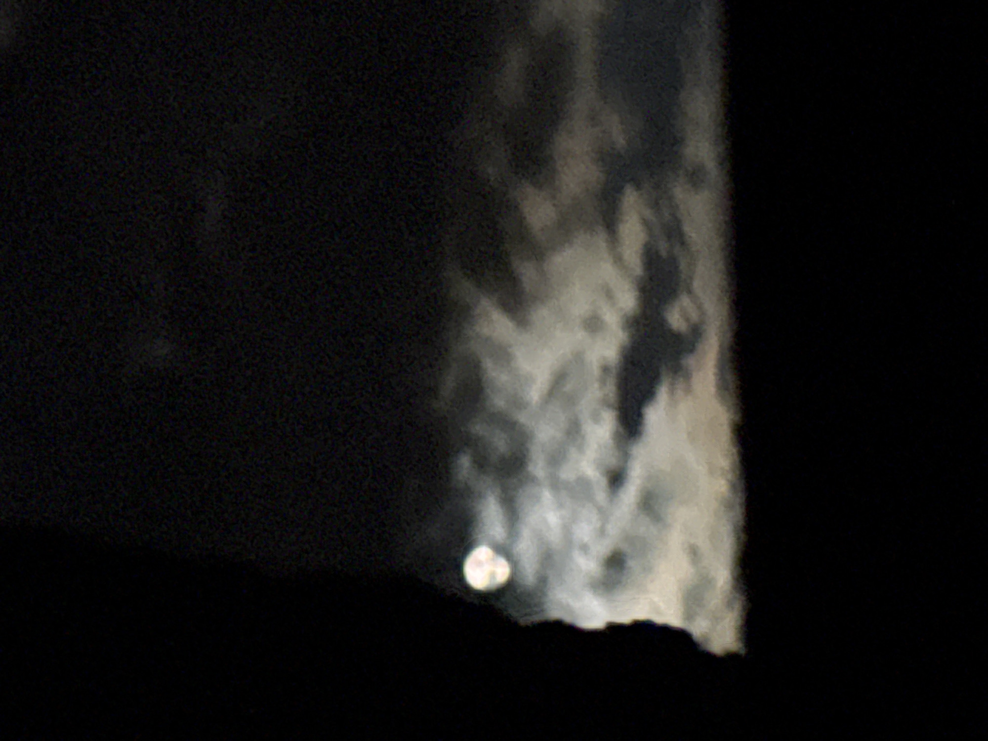 The image presents a nocturnal scene featuring a celestial body, likely the moon, partially obscured by both clouds and a dark, silhouetted landmass. The dominant subject is a bright, circular, and somewhat blurry light source positioned in the lower-center, emitting a soft glow and appearing to rise above a dark, undulating silhouette that spans the bottom of the frame, suggestive of mountains or hills.
The sky is largely dark, with the left side being almost entirely black and textured with visual noise, indicating extremely low light conditions. The right half of the image is filled with dynamic, illuminated clouds, showing varying shades of gray and white, suggesting their texture and movement against the dark background. These clouds are diffuse and appear atmospheric, possibly contributing to the slight haziness around the bright celestial object. No specific human figures or man-made objects are visible, nor is any text present. The overall setting is an outdoor, natural landscape at night, with the prominent details being the interaction of light from the celestial body with the clouds and the landforms below.