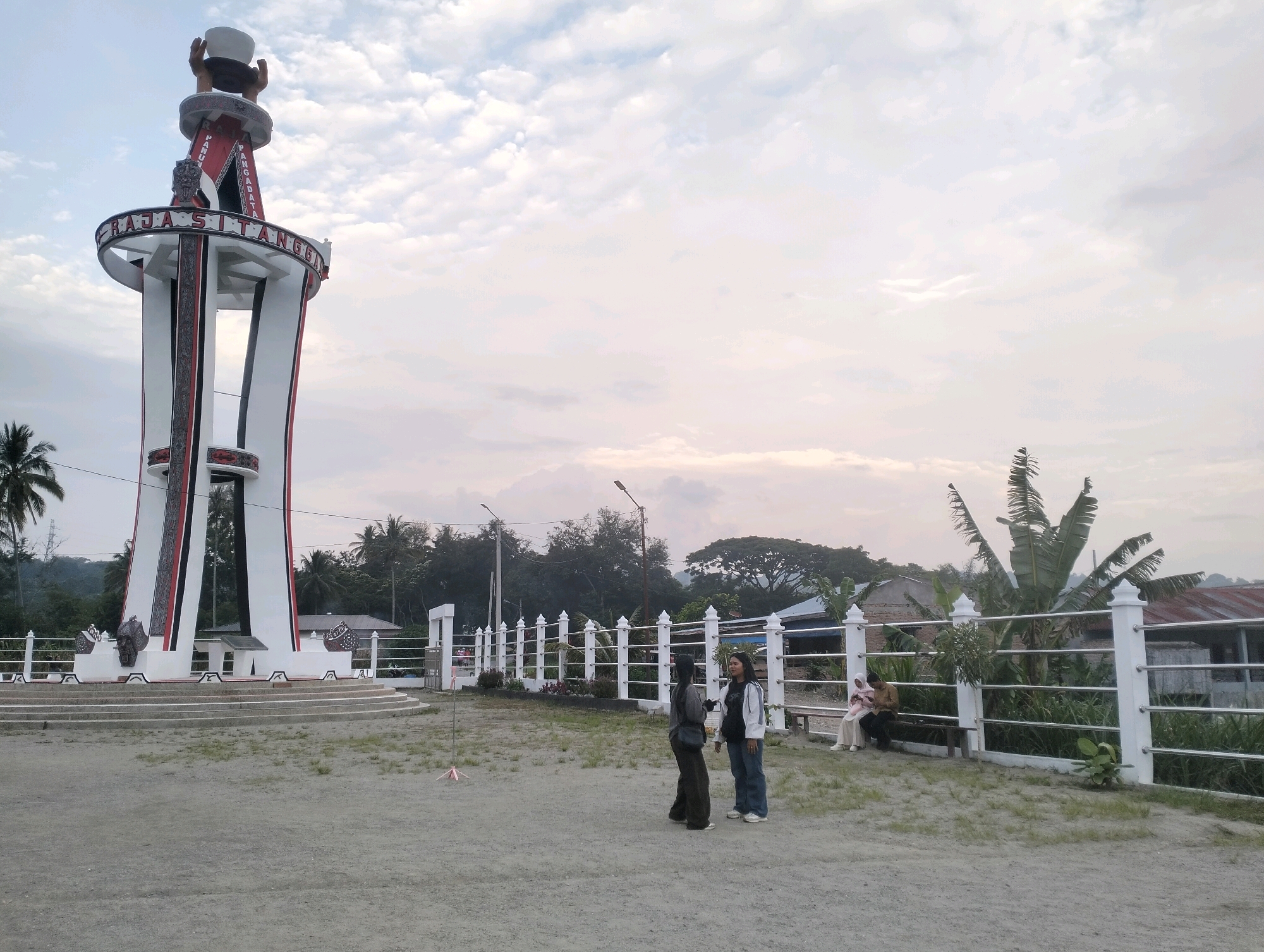 The image shows a large monument, possibly a tower, in the city of Parlondut, Indonesia. The monument is white with red and black accents and has text around its upper section. Two women are standing in the foreground, talking to each other. In the background, there are trees, buildings, and a white fence. A few other people are visible sitting on a bench near the fence. The sky is cloudy.