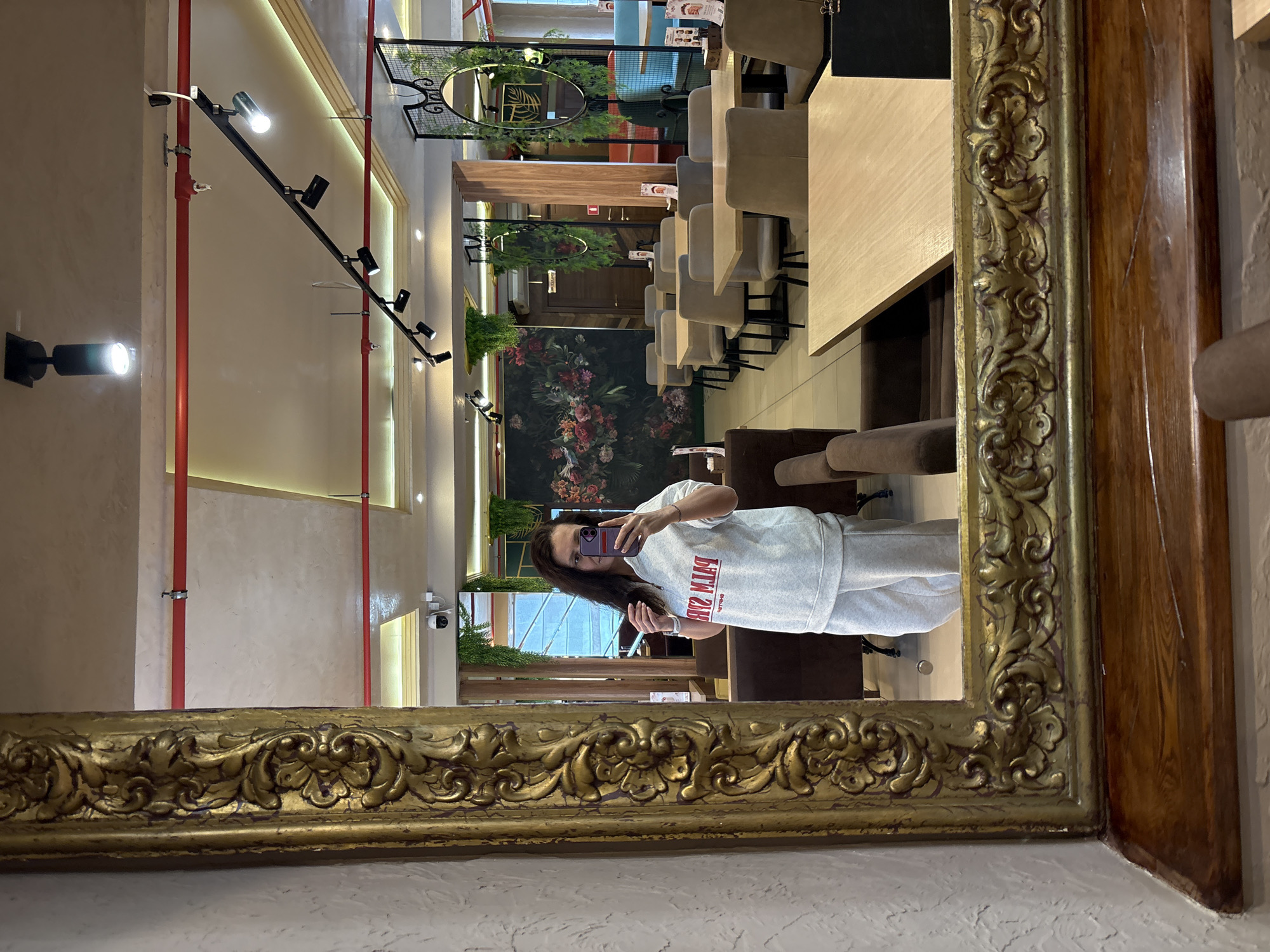The image captures a mirror selfie taken by a woman in what appears to be a cafe or restaurant interior. The large mirror, which occupies the foreground and right side of the frame, has an elaborate, ornate gold frame with a distressed finish, and a wooden panel adjoins it on the right.
The central subject is a woman, reflected in the mirror, holding a smartphone with a purple case up to take the photo. She has long, dark hair and is wearing a light grey long-sleeved top and matching light grey sweatpants. Visible text on her shirt, reversed, appears to read "PINK SUM". Her expression is neutral as she focuses on the phone.
The reflected interior of the establishment shows a modern and somewhat eclectic decor. Along the left wall, exposed red pipes run vertically, suggesting an industrial aesthetic, alongside track lighting with multiple spotlights illuminating the space. Further back, a large, dark floral mural or wallpaper featuring vibrant flowers and greenery covers a section of the wall. In front of this, there are dark upholstered banquette seats and numerous light-colored tables paired with grey or beige upholstered dining chairs. More seating areas and tables are visible further into the room. Decorative elements include hanging green plants and small round mirrors with foliage. A security camera is mounted on a wall in the upper left of the reflection. The overall lighting is bright, indicating the establishment is open, likely during the day or evening. The context provided indicates the location is Neftekamsk, Russia.