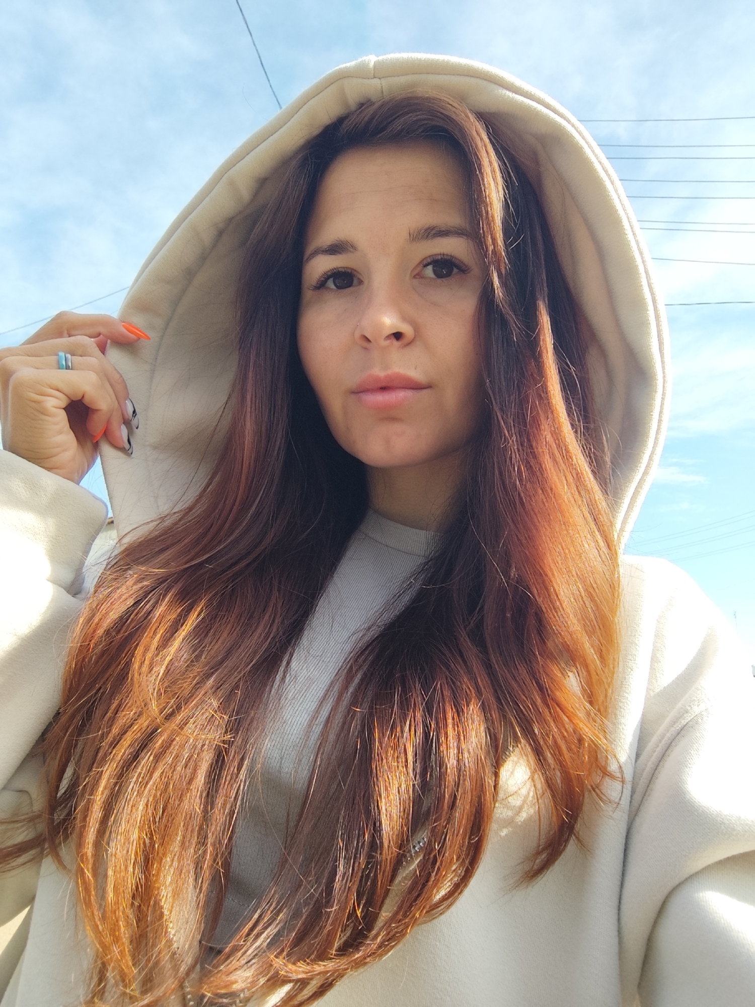 A woman with long auburn hair, wearing a light-colored hooded sweatshirt, poses for a selfie outdoors. She is wearing two rings on her left hand and her fingernails are painted with a combination of orange and black/white patterns. A section of blue sky is visible in the background with some power lines running across the top right of the frame. There is no definitive information to confirm the location is in Фоки, Russia.