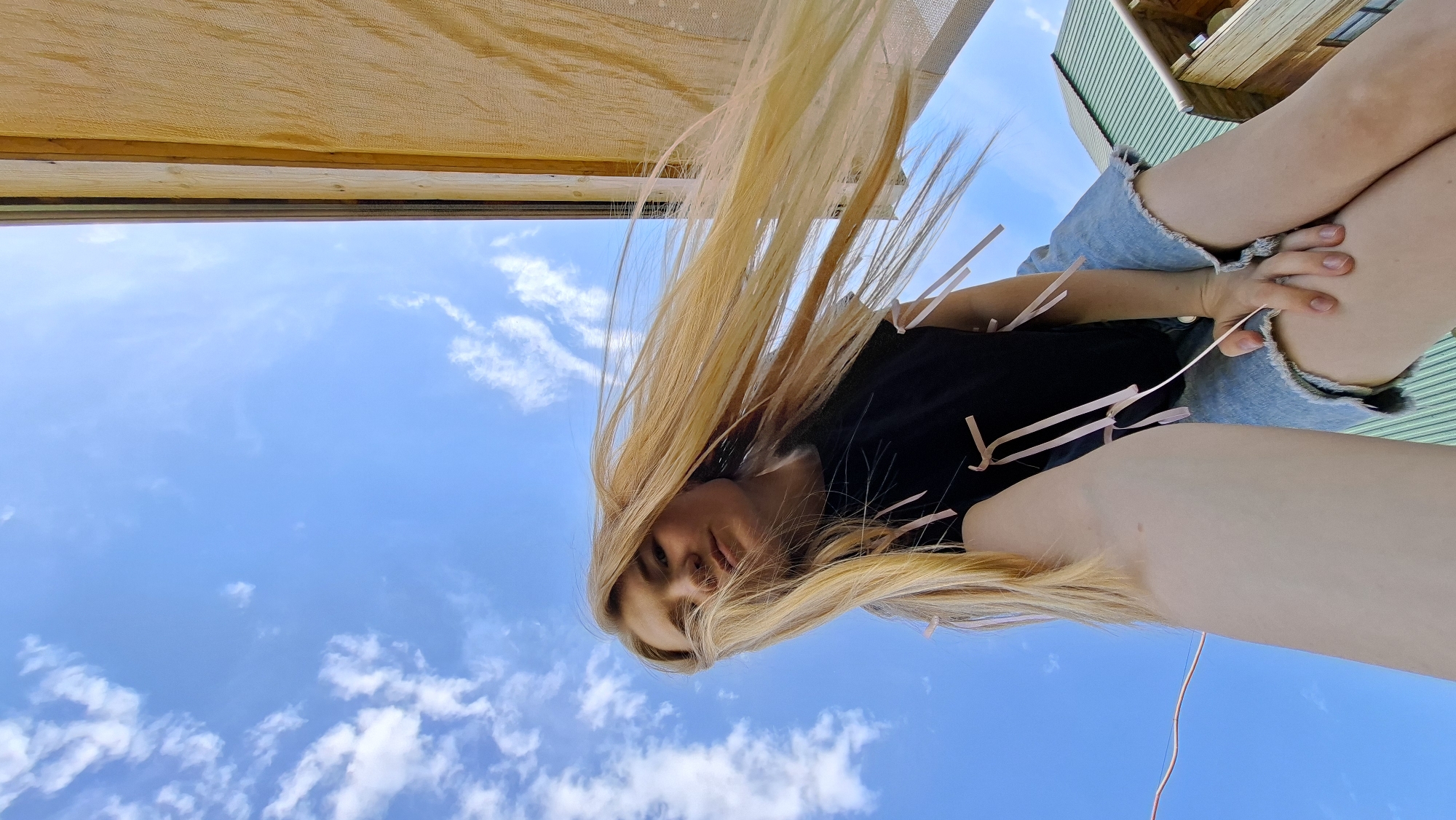 A low-angle shot looking up at a fair-skinned woman with long blonde hair. She's wearing a black shirt and blue denim shorts. Pink streamers are attached to her shirt. Above her is a blue sky with scattered white clouds. At the top edge of the photo, there is what appears to be a wooden structure with beige fabric. The location is Большой Букор, Russia.