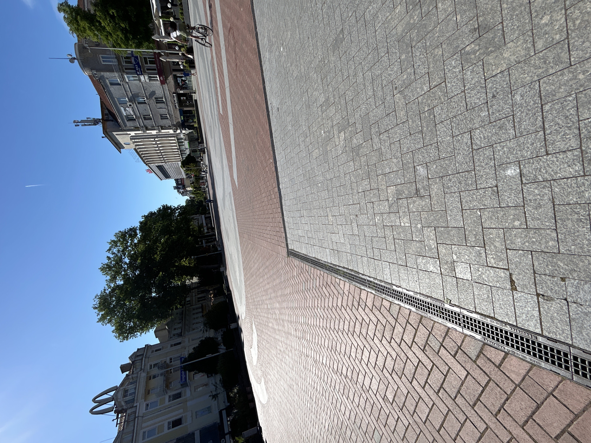 The image shows a section of a pedestrian zone in Varna, Bulgaria. The area is paved with two types of stone. A gray, rectangular cobblestone pattern covers the right portion of the image. A brick-colored paving stone is laid in a herringbone pattern to the left. A metallic drainage grate runs lengthwise between the two paved sections. Buildings and trees line the left side of the image, under a clear blue sky. A bicycle and pedestrians can be seen in the upper left.