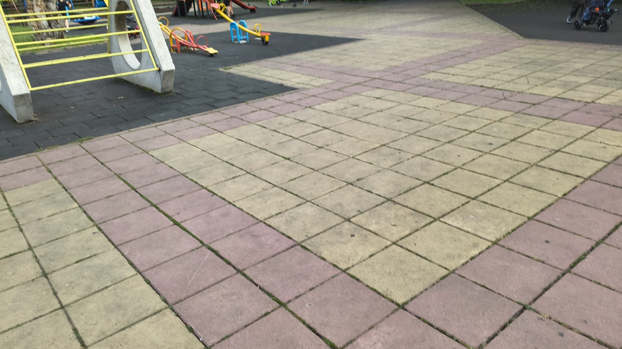 The image depicts an outdoor playground area located in Shumen, Bulgaria. The foreground and midground are predominantly covered by a paved surface composed of square concrete tiles. These tiles are arranged in an alternating pattern of faded light beige and muted reddish-pink, with green grass or moss visible growing within the grout lines.
To the left, a section of dark grey, possibly rubberized, safety flooring is visible beneath playground equipment. This equipment includes the concrete support structures and horizontal yellow metal rails of what appears to be a climbing frame or slide. Adjacent to this, also on the dark matting, are two seesaws: one with a red base and yellow beam, and another with a blue base and yellow beam. Partial views of other red playground structures are also visible in this area.
In the background, beyond the playground equipment, green foliage from trees and grass is present. On the far right of the image, the lower half of an individual pushing a dark-colored stroller, which contains a child, is visible on a paved path. Further into the background on the left, several indistinct figures are partially obscured by trees, suggesting other people are present in the park environment. The scene is illuminated by diffused daylight.