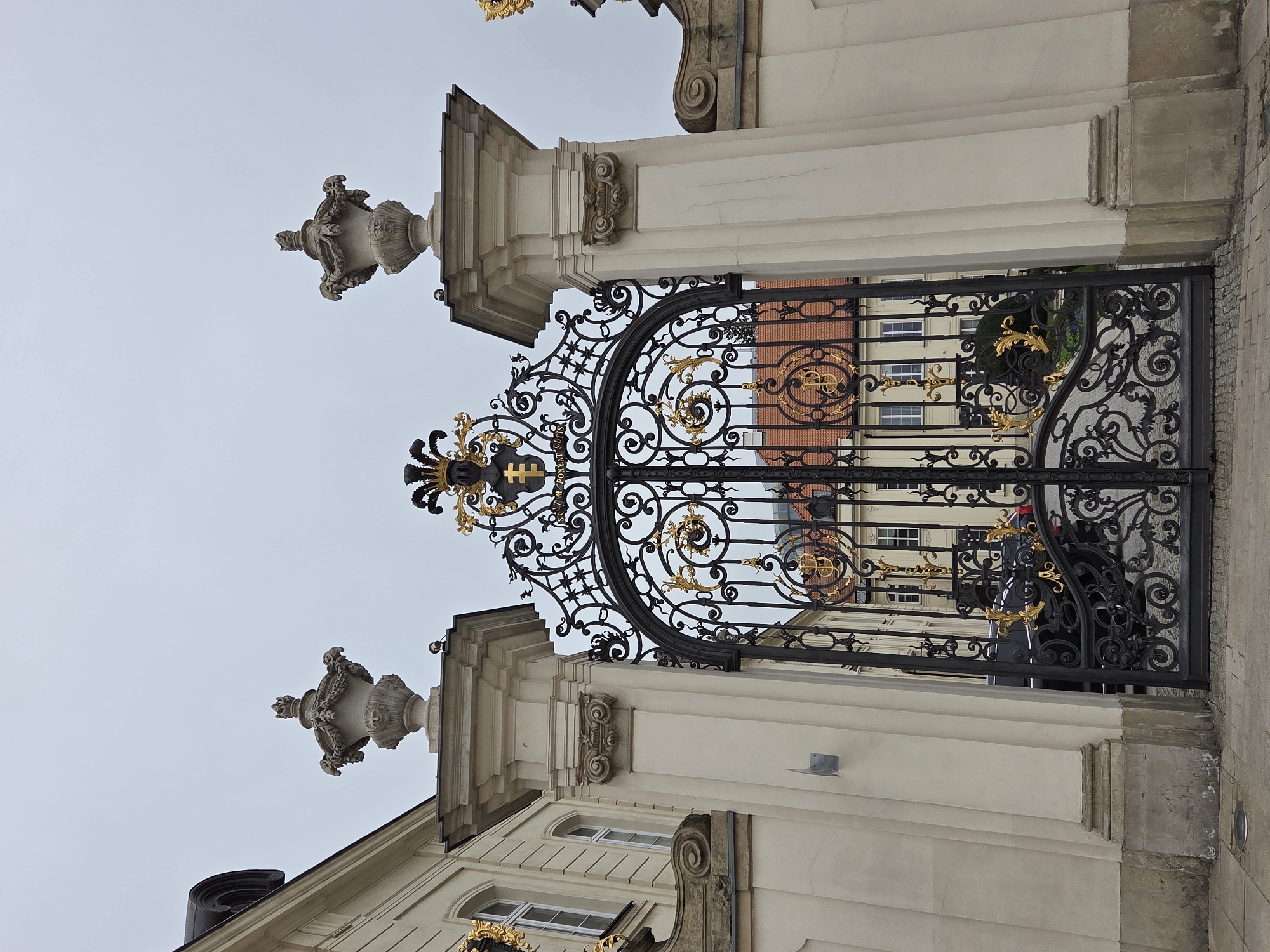 The image depicts a wrought iron gate in Warsaw, Poland. The gate is predominantly black, adorned with gold embellishments. It stands between two white, classically styled pillars topped with decorative urns. The gate's design is intricate, featuring scrolls and curves. Through the gate, a building with a red roof and white facade is visible. The sky is overcast.