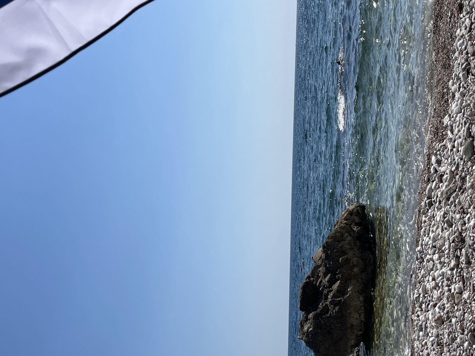 The image shows a sunny day at a pebble beach with clear blue water. A large rock is partially submerged in the shallow water near the shore. The sea extends to the horizon under a cloudless blue sky. The foreground is dominated by a dense collection of smooth, grey and white pebbles. In the upper right corner, a portion of a white and blue striped beach umbrella is visible. There appear to be a few distant figures in the water, possibly swimming or sunbathing. The scene is located at the Queen's Beach (Kraljičina plaža) in Bar, Montenegro, a well-known pebble beach area.