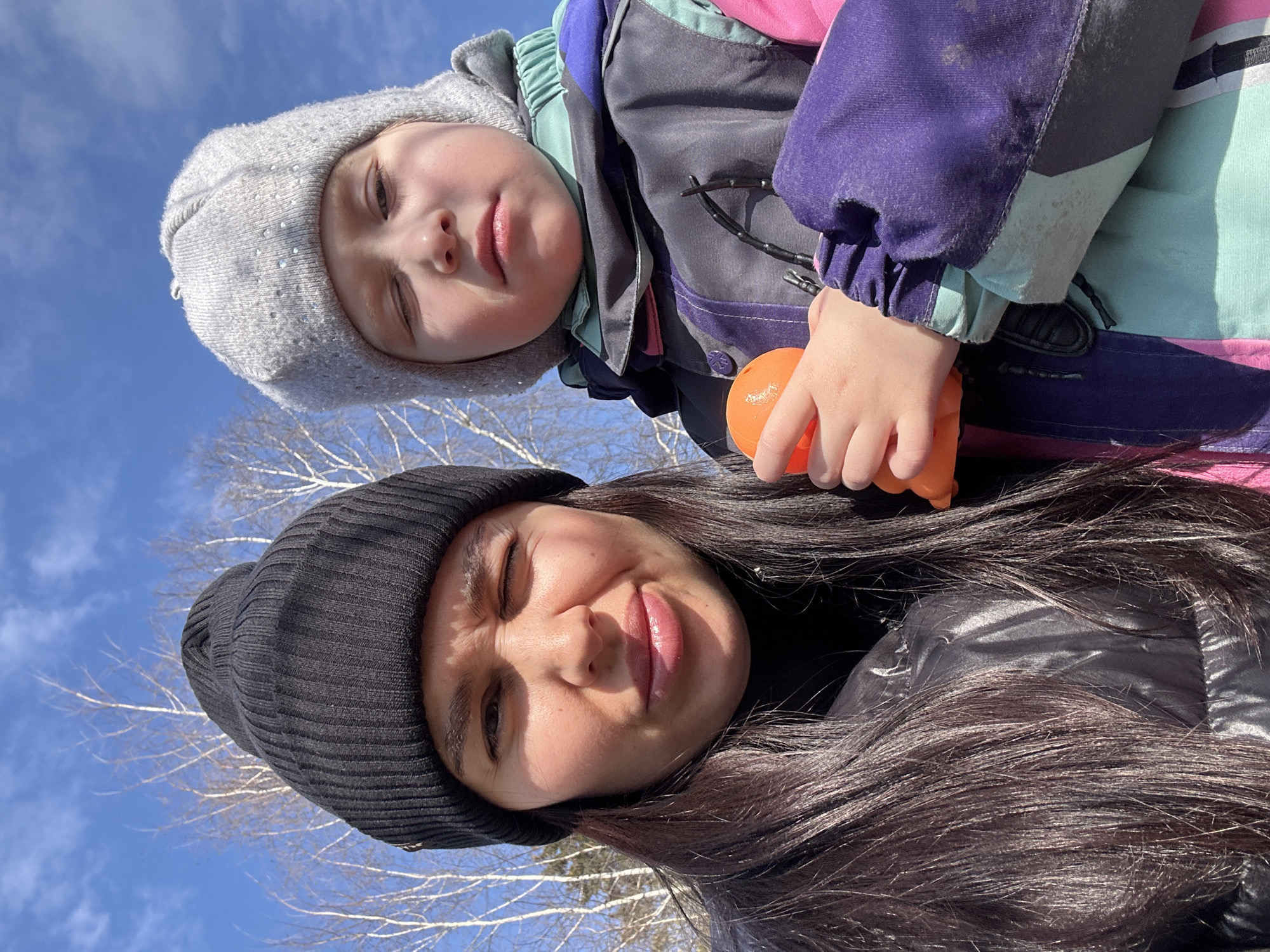 The image shows a selfie taken outdoors on a sunny day in a location with trees, potentially in the city of Chaikovsky, Russia. A woman with long dark hair wearing a black beanie is positioned below and slightly to the right in the frame. Above her is a young child wearing a gray beanie and a colorful winter jacket; the jacket features purple, gray, and turquoise panels. The child is holding a small orange object in their hand. The sky is blue and visible in the background. The image is oriented at an unusual angle.