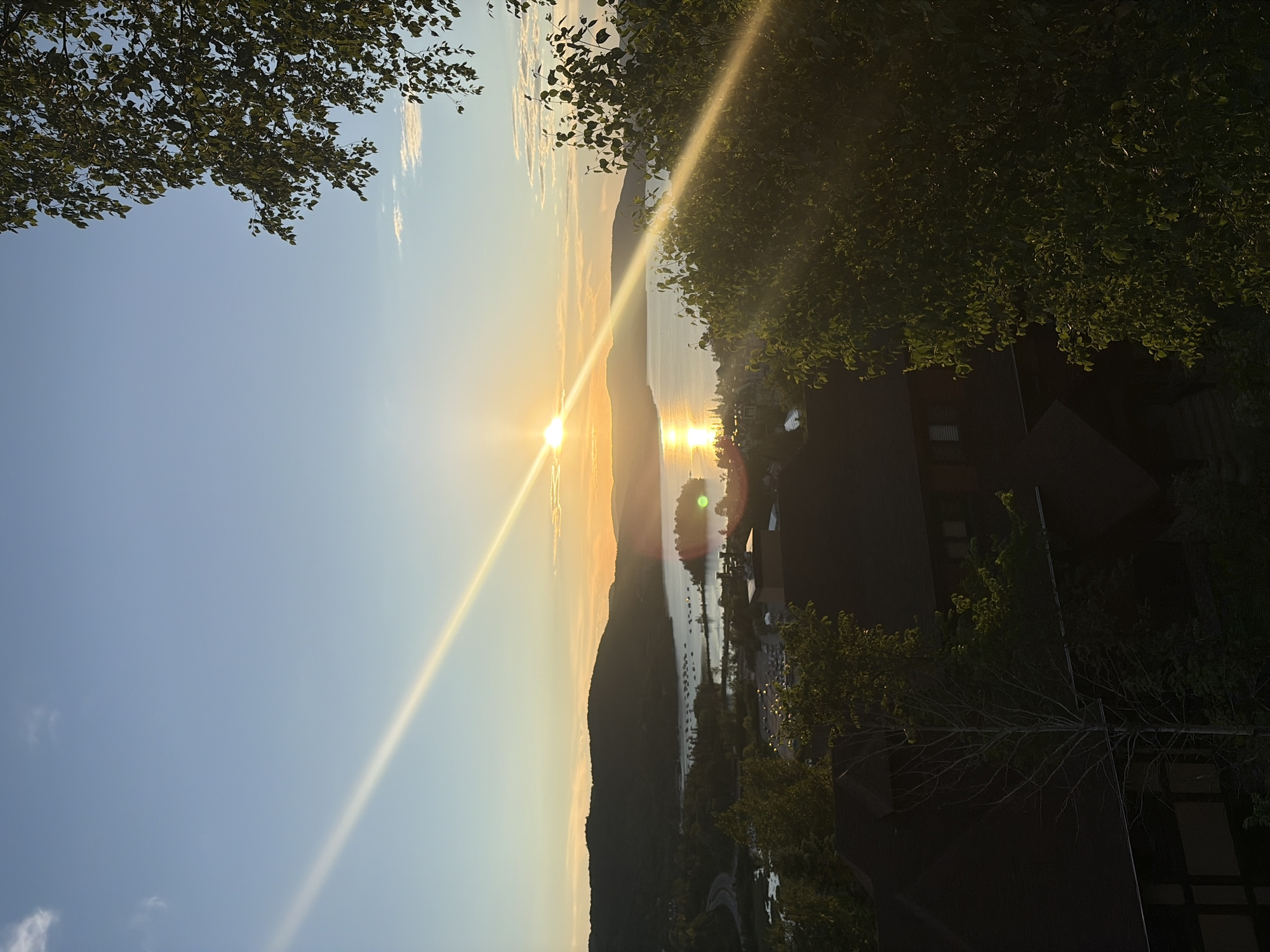 This is a high-angle, full-shot, vertical photograph of a sunset over a body of water in Mont-Tremblant, Canada. The sun is setting on the horizon, casting a bright glare across the water and a diagonal beam of light across the sky. In the foreground, there are trees and the dark roofs of buildings. The middle ground shows a bay with boats moored, and beyond that, rolling hills covered in trees. The sky is clear with a few small clouds. The overall impression is of a serene and picturesque landscape at dusk.