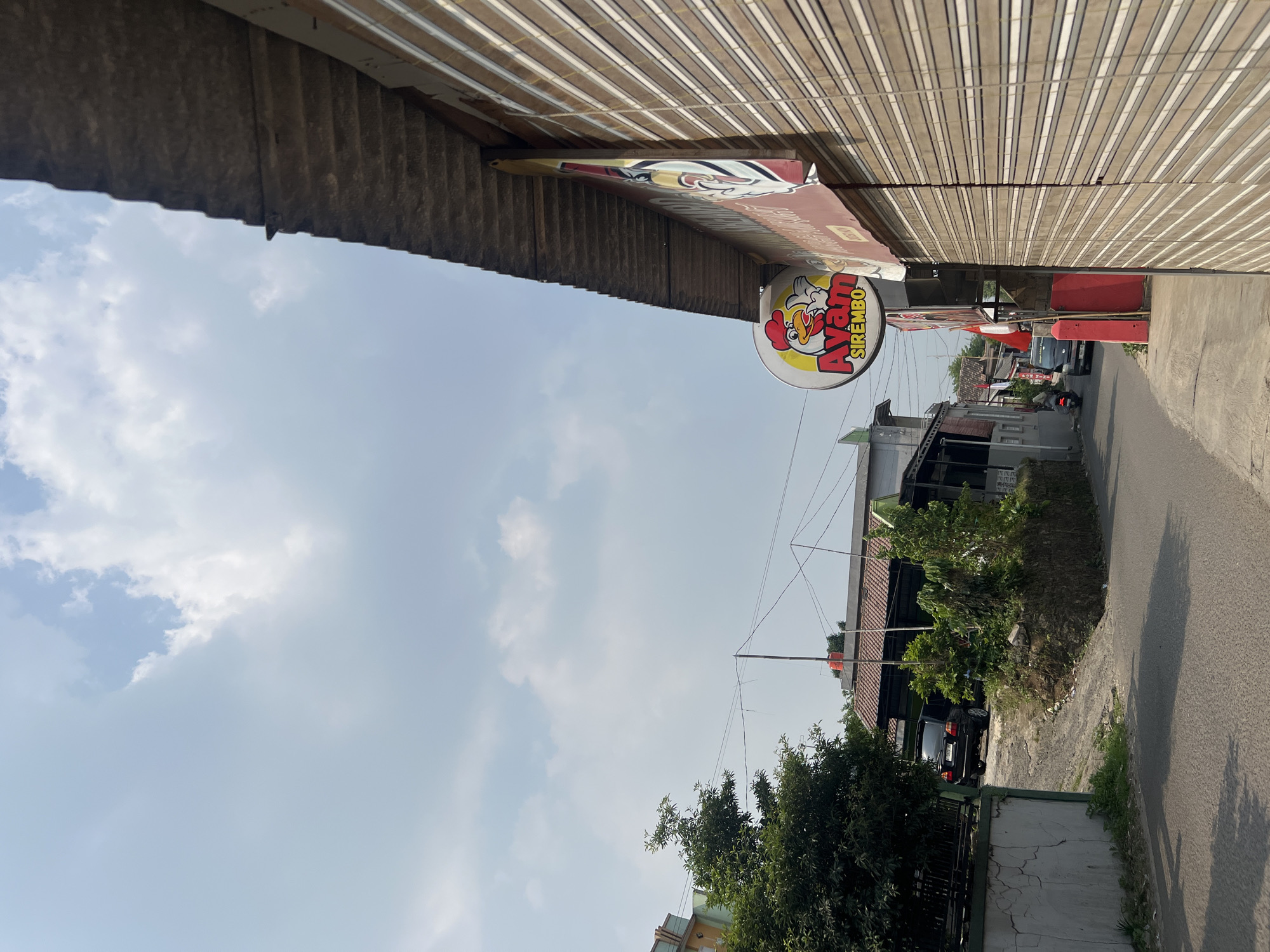 The image depicts a street scene in Klapanunggal, Indonesia, with a focus on a commercial establishment. On the right side of the frame, a building features a closed bamboo-style shutter and a prominent circular sign with a cartoon chicken and the text "Ayam SIREMBO" in red and yellow. Adjacent to this is a larger rectangular sign, partially visible, with red and white lettering. The building's roof is weathered and made of corrugated material.
The street itself is paved and curves to the left in the background. To the left of the street, there is a grey, cracked wall and a lush green tree. Further back, several buildings with tiled roofs are visible, including one with a visible red water tank on top. A black SUV is parked on a dirt incline leading up to one of these buildings. A motorcycle is in motion on the street, ridden by a person wearing a helmet. The sky is mostly blue with scattered white clouds. The overall impression is of a quiet, possibly residential or commercial street on a clear day.