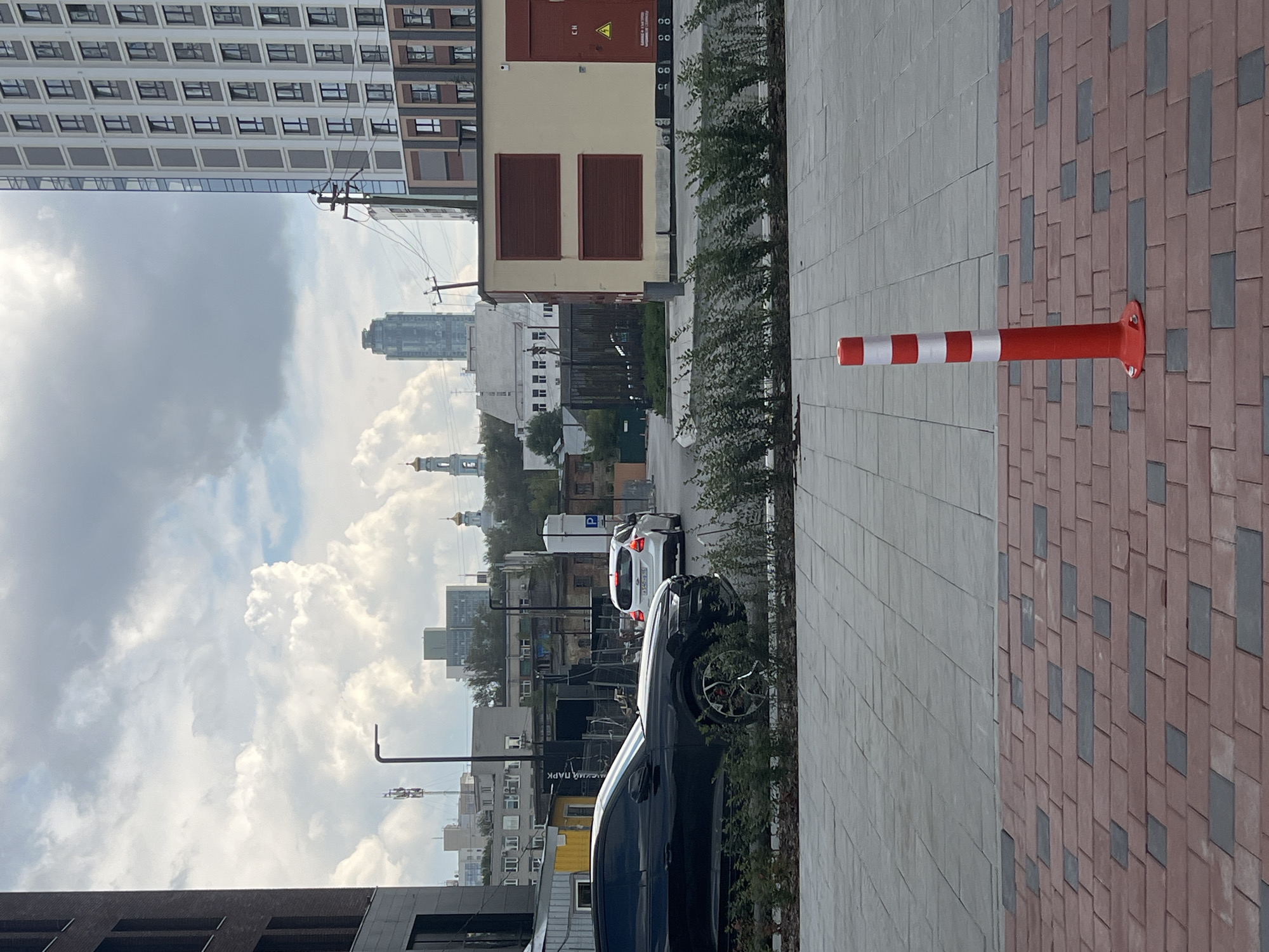 The image depicts an urban scene in Yekaterinburg, Russia, with a focus on a modern building facade and a street. In the foreground, a red and white striped traffic bollard stands on a patterned brick sidewalk. Behind the bollard, a gray paved surface leads to a row of green bushes. To the left, a black car is parked. Further in the background, various buildings of different architectural styles are visible, including a tall, modern residential building with many windows on the right, and smaller, older structures. A tall, slender skyscraper is visible in the distance. In the sky, white and gray clouds are present. The overall impression is of a developing urban area.