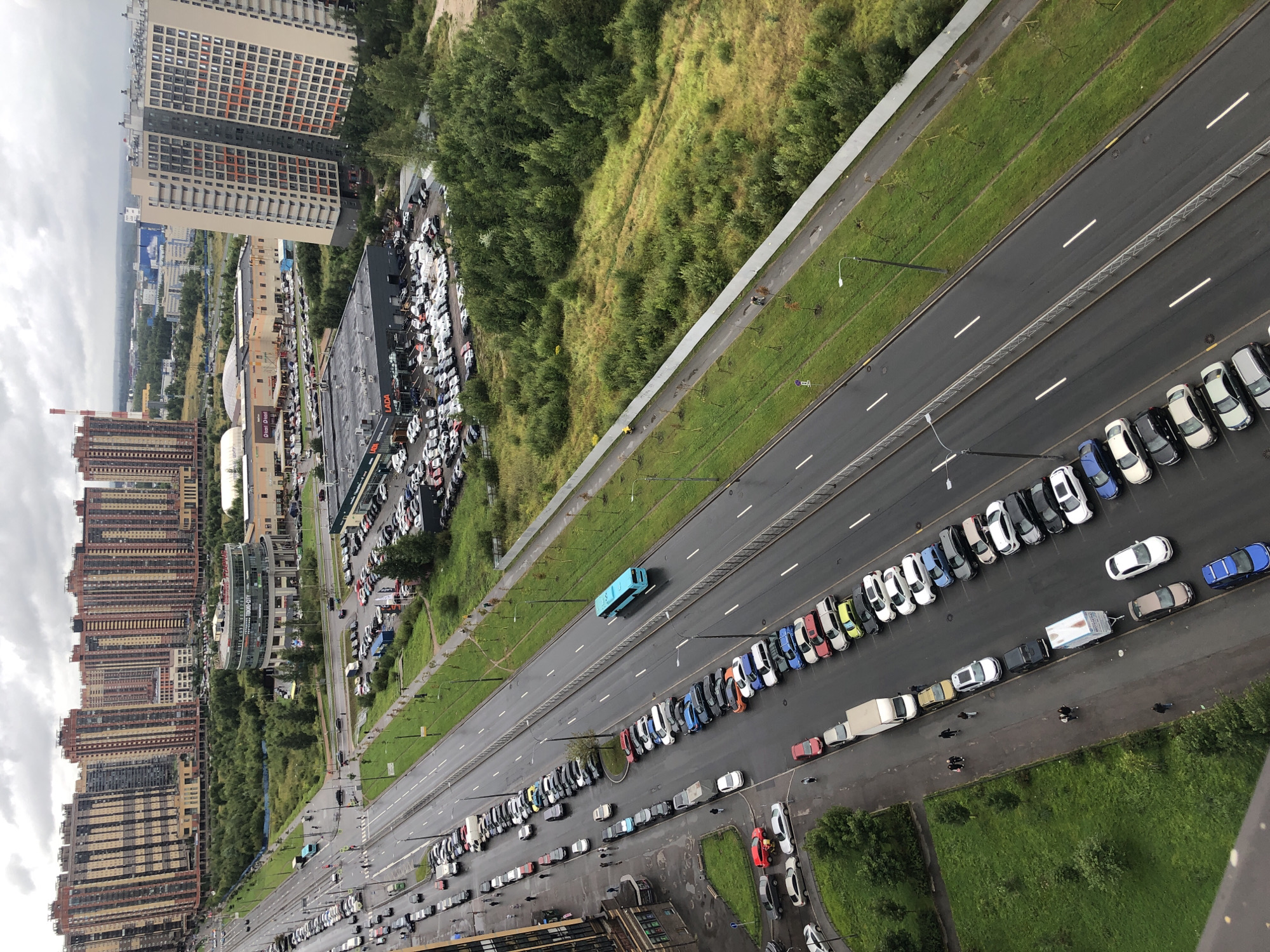 This aerial view captures a wet, overcast urban scene. A multi-lane road, bordered by parked cars and a grassy embankment, dominates the foreground. A bright blue bus travels down the road. In the background, a cluster of tall, modern residential buildings with patterned facades stands out against the sky, interspersed with smaller commercial buildings and a large parking lot filled with vehicles. Green trees and patches of grass surround the buildings, indicating a mix of residential and commercial development with green spaces.