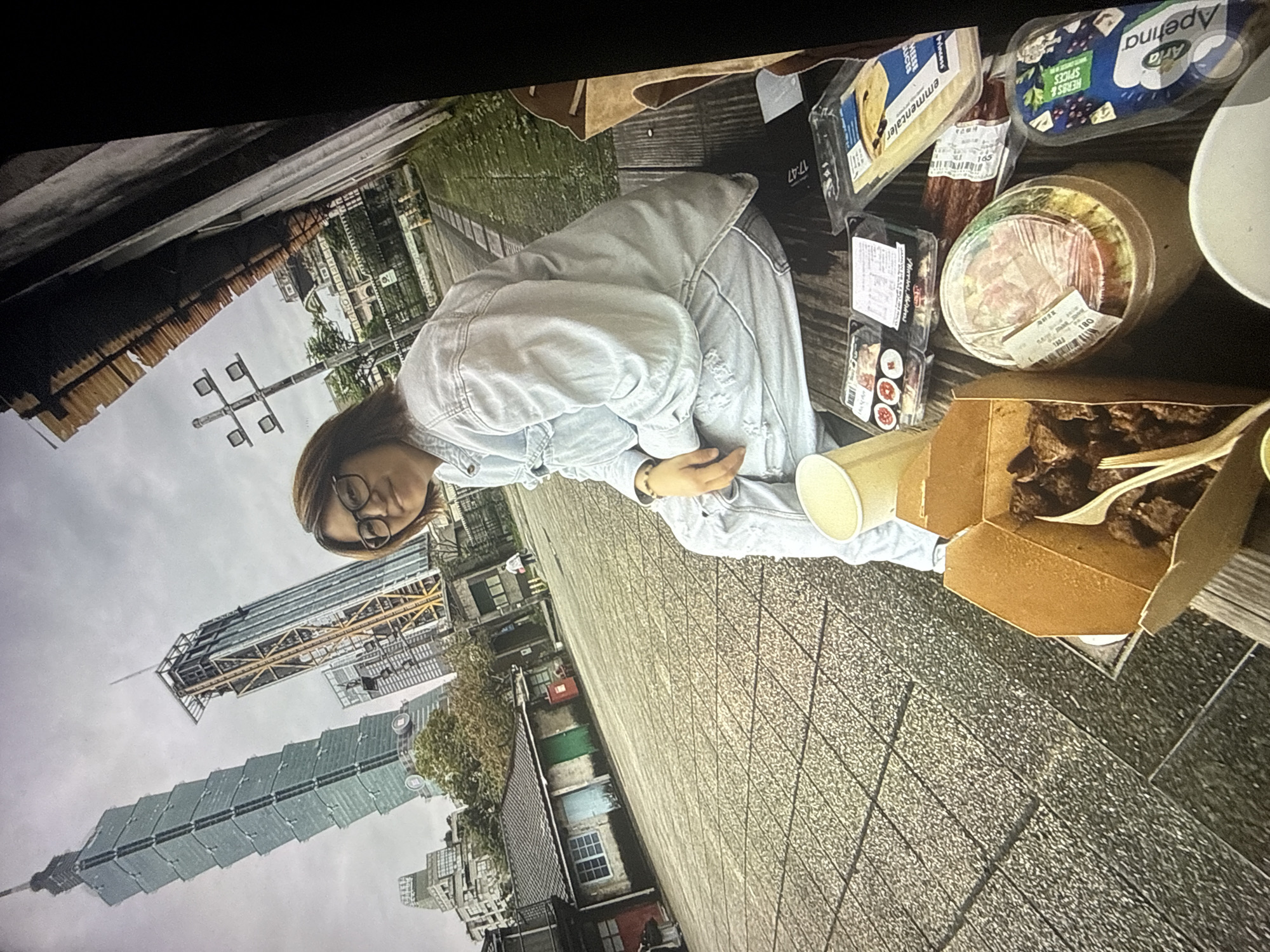 A young woman with dark hair and glasses sits on a wooden bench outdoors in Taipei, Taiwan, with a picnic spread in front of her. She is wearing a light blue denim jacket and distressed jeans. The background features the iconic Taipei 101 skyscraper, along with other urban buildings and greenery. The picnic includes a takeout container of food, sushi, a salad, cheese slices, and a package of herbs and spices. The ground is paved with stone tiles, and a portion of a building wall is visible to the right. The sky is overcast.