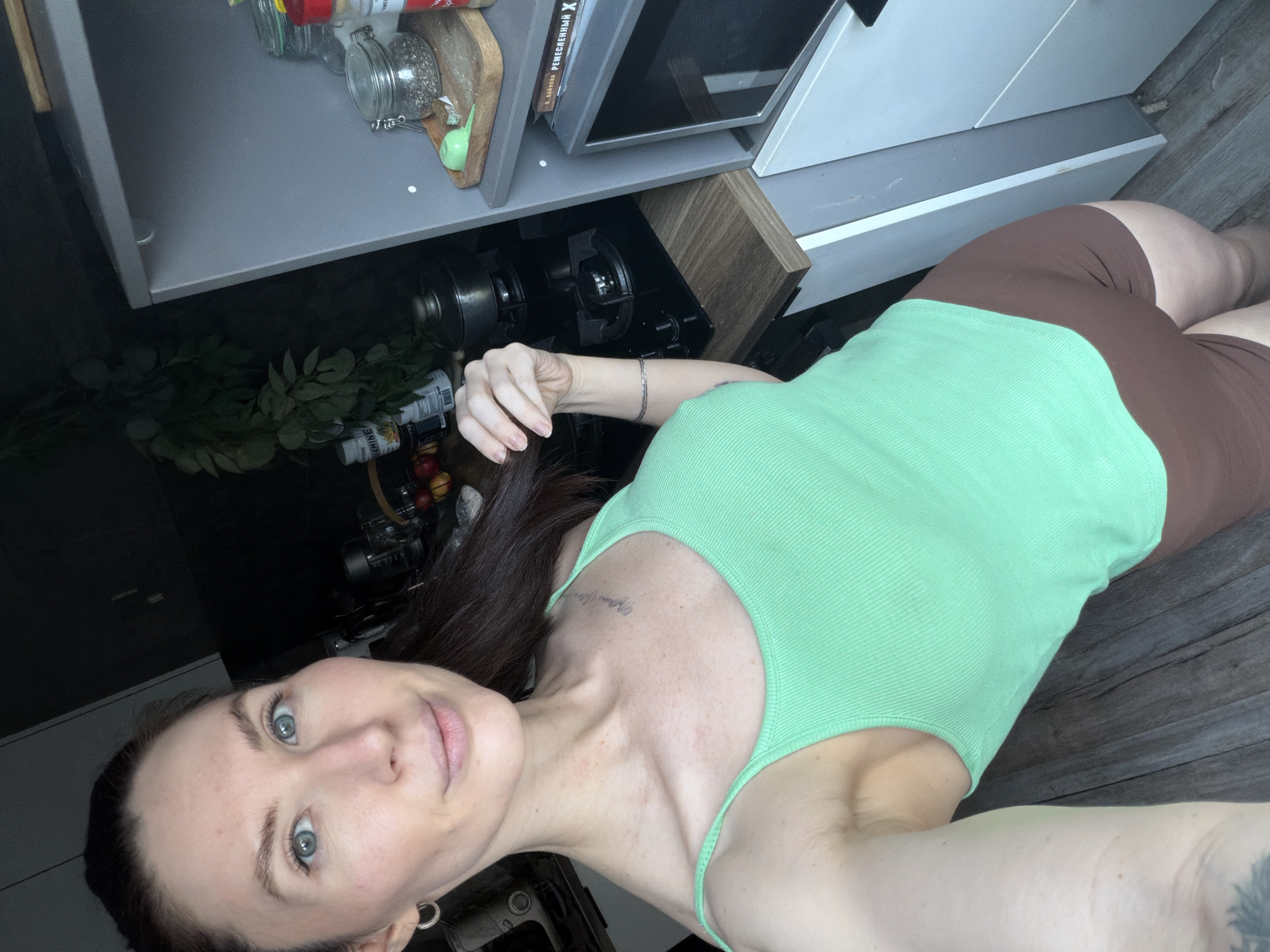 The image is a selfie of a woman taken from a low angle, appearing to be lying on a dark wooden floor and looking upwards. The setting is indoors, resembling a modern kitchen with contemporary cabinetry and appliances visible above her.
The subject is a woman with long, dark brown hair, which is pulled back. She has light blue-green eyes and a fair complexion, with subtle freckles visible on her décolletage. She wears a fitted, ribbed light green tank top and brown cycling shorts. A slender silver chain bracelet is on her right wrist, and a small silver hoop earring is in her left ear. A script tattoo is visible on her right collarbone area. She is looking directly at the camera with a neutral to slightly serene expression.
Above her, various kitchen elements are arranged. A light grey cabinet unit is mounted, housing a silver microwave oven. Below the cabinet, a black induction cooktop is integrated into a counter. On the cabinet's shelf, several items are visible, including glass jars (one containing black peppercorns), various bottles, a wooden cutting board, and a small green utensil. Artificial or dried dark green foliage is placed in the background. A book with a red spine is also on the shelf, with the Russian word "РЕЦЕПТЫ" (Recipes) and the letter "X" partially visible on its spine. The lighting appears to be natural daylight. The image's location context is Perm, Russia.