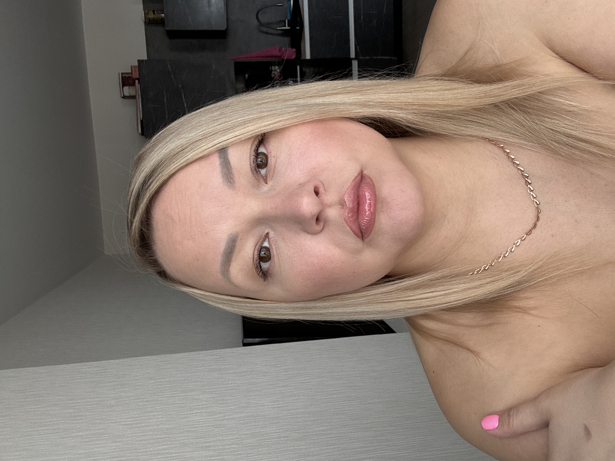 A blonde-haired woman with a gold chain around her neck poses indoors. The location appears to be a room with gray walls and a black and gray cabinet in the background. A finger with a pink painted nail is visible in the lower right corner. The city and country of origin are not discernible from the image.