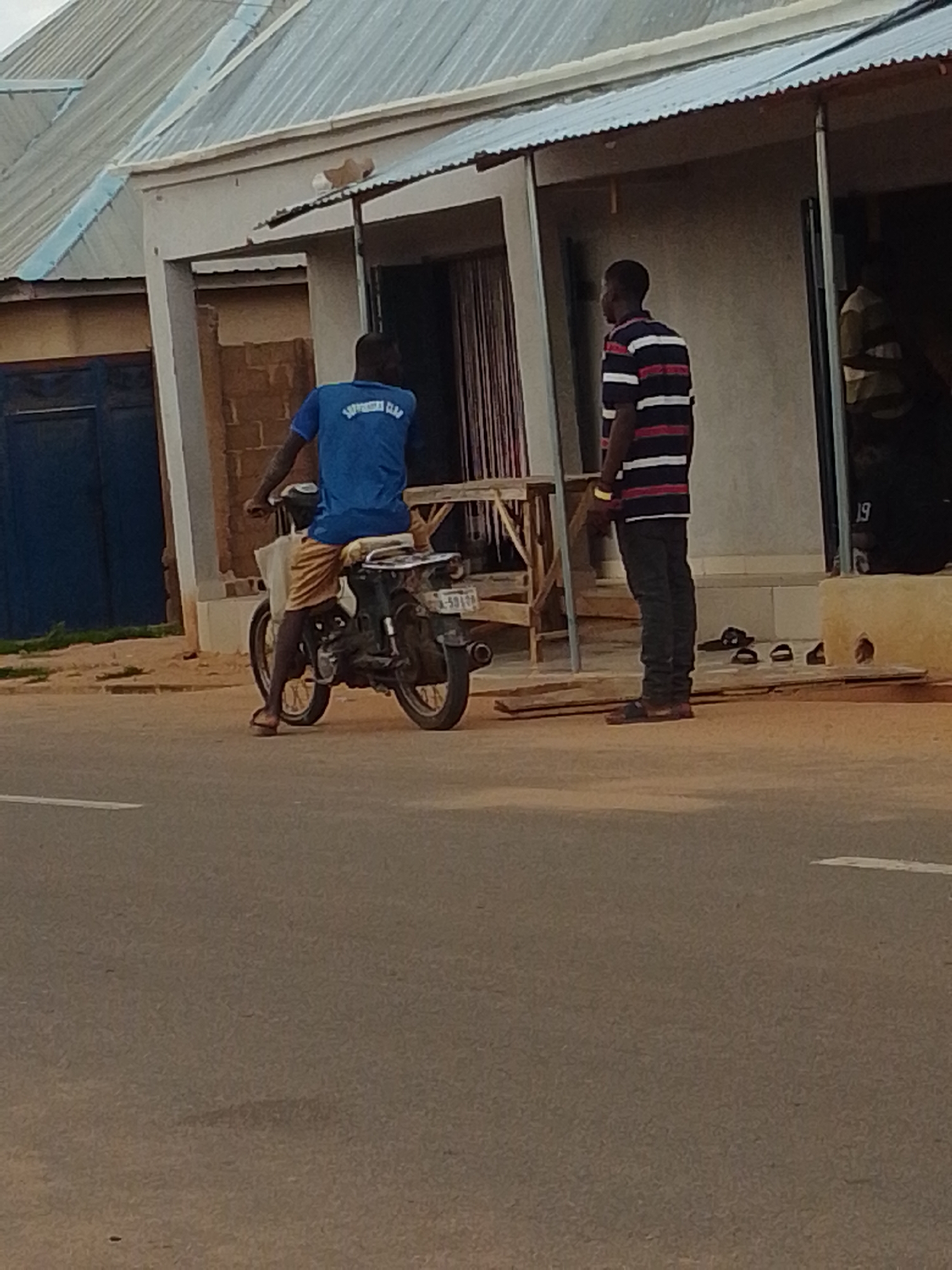 The image captures an outdoor scene on a street in Tempure, Nigeria. Two men are present. In the foreground, a man wearing a blue t-shirt with "SOUTHERN CLUB" visible on the back, and tan shorts, is seated on a motorcycle. The motorcycle is dark in color, and the man is holding onto the handlebars. Behind him, further down the street, stands another man wearing a horizontally striped t-shirt in navy, red, and white, and dark trousers. He appears to be facing away from the motorcycle rider, towards a building. The building behind them has a light-colored facade with a corrugated metal roof. A blue door is visible on the left side of the building. The ground is paved, and there is a white line marking on the road in the foreground. Various objects, including what appear to be sandals, are scattered on the ground near the building's entrance. The overall lighting suggests it is daytime, possibly late afternoon.