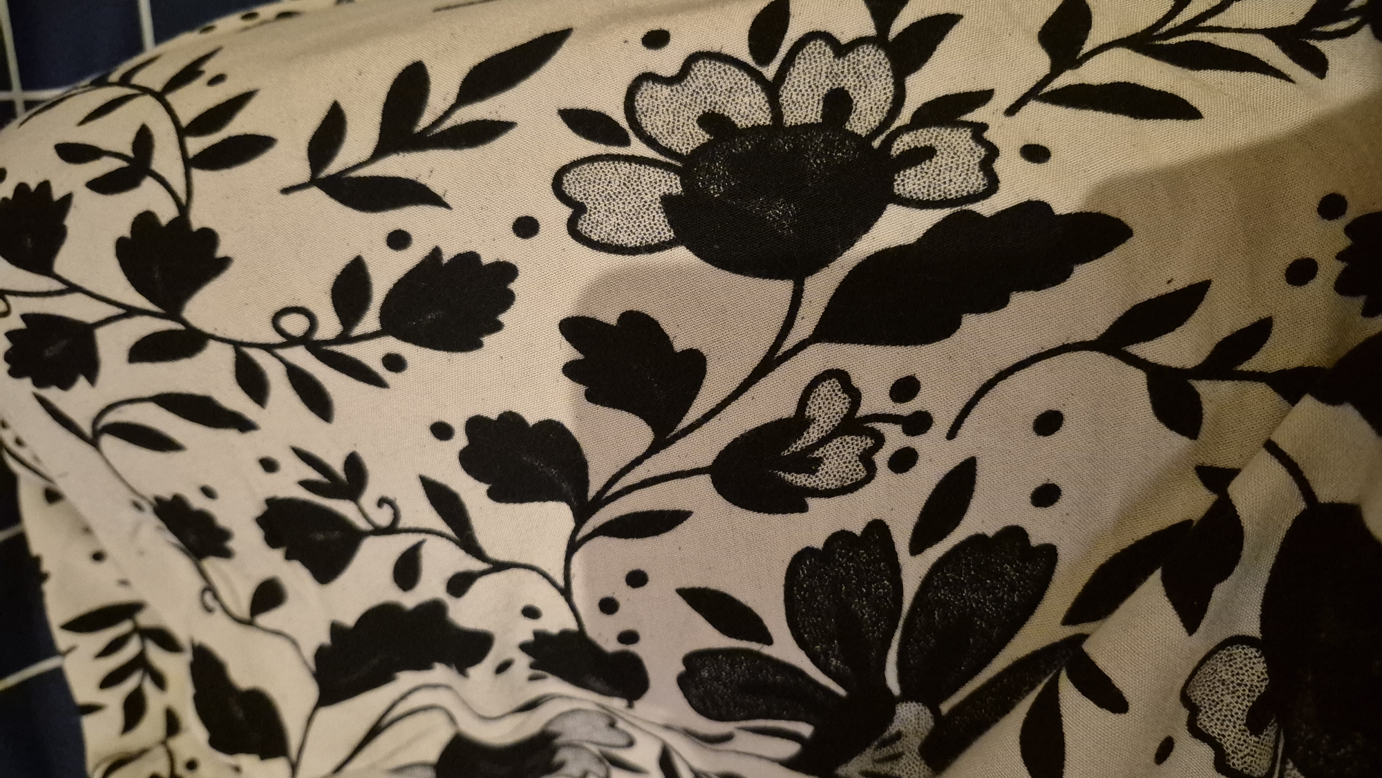 The image shows a close-up of a cream-colored fabric with a black floral pattern. The fabric is draped and creased, suggesting it might be clothing or bedding. The floral design features stylized flowers with multiple petals and delicate stems with leaves and small dots. Some of the flower petals have a textured, stippled pattern within them, creating a contrast with the solid black outlines. The background appears to be a dark blue and white striped material, possibly another piece of fabric or a surface. The lighting is dim, casting soft shadows on the fabric, and the overall impression is one of detail and texture. The image does not contain any elements that would allow for specific identification of the location in South Tangerang, Indonesia.