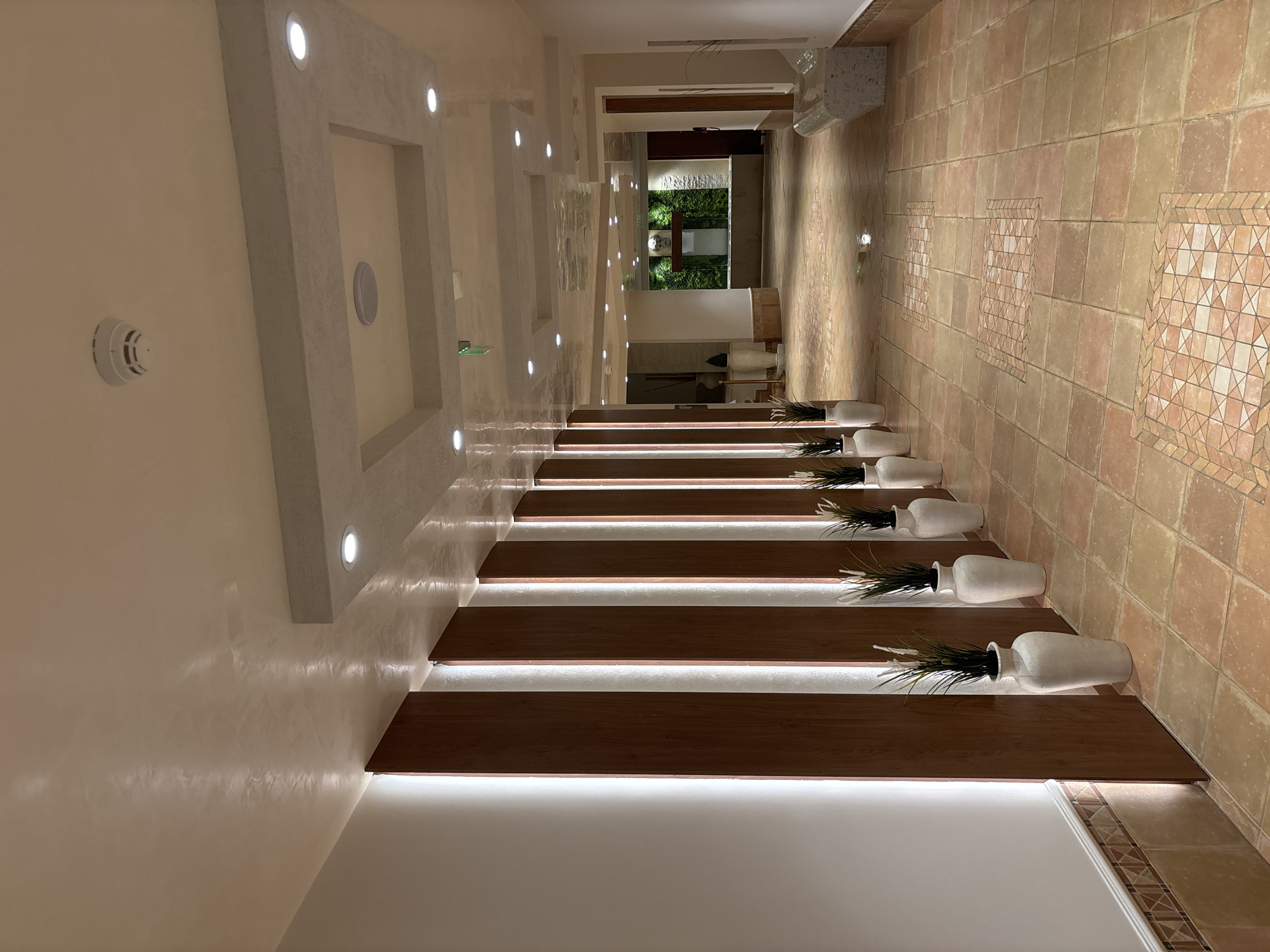 This is a photograph of an interior hallway with a tiled floor and a ceiling with recessed lighting. The hallway walls are lined with vertical wooden panels interspersed with vertical light fixtures. Several white vases with decorative grass are placed along the wall. In the distance, a green wall covered in foliage is visible, with a sculpture above it. The floor is covered with terracotta-colored square tiles, with patterned tile inserts. A stone bench is visible on the right side of the hallway. The scene appears to be a hotel corridor or a similar public indoor space. The city of Razliv, Bulgaria, is not mentioned or depicted in the image itself, and there is no information within the image to suggest this specific location.