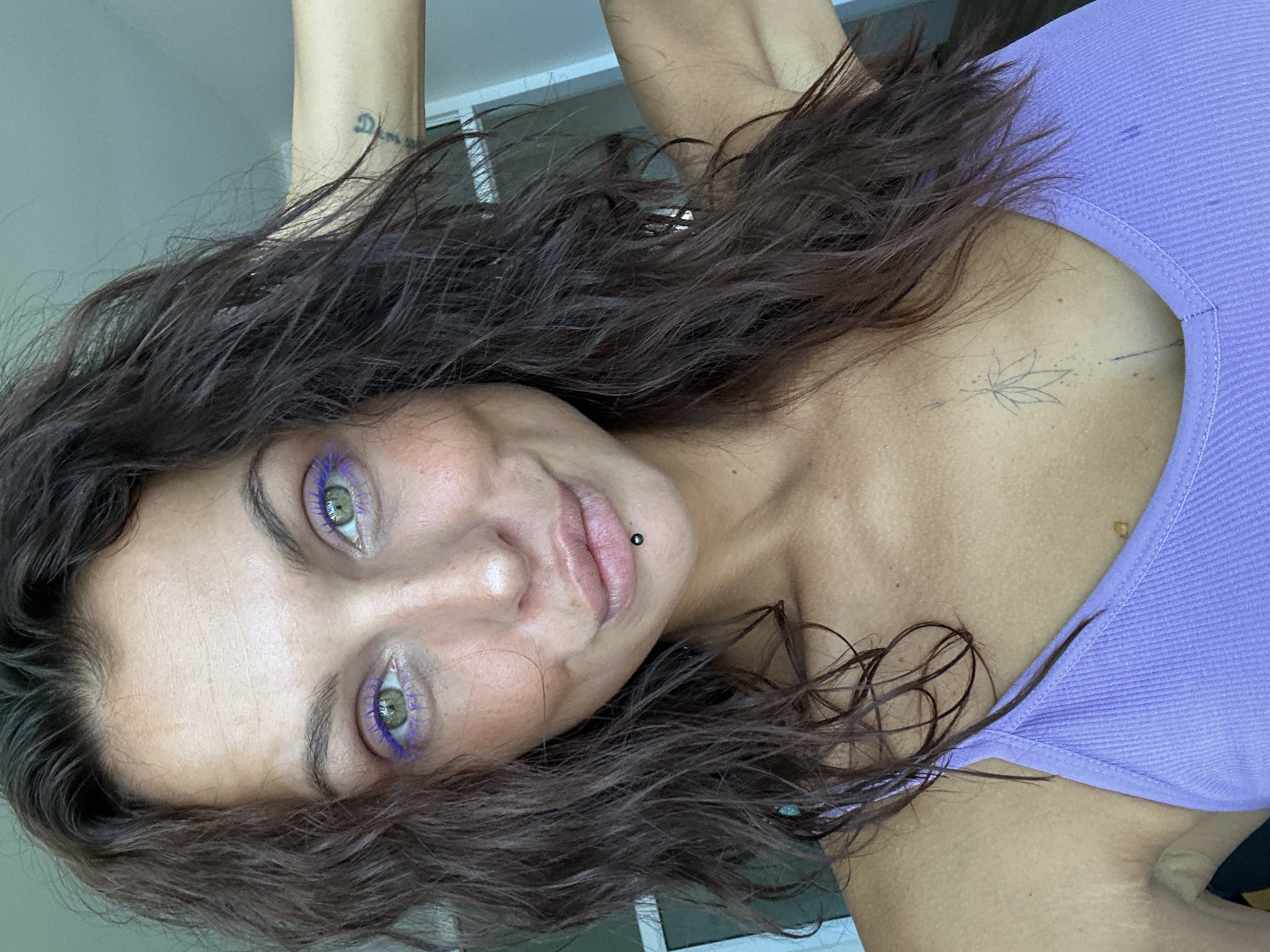 A young woman with dark, wavy hair is shown from the chest up. She is wearing a purple tank top. She has green eyes with purple mascara. Her lips are slightly parted, and she has a black stud piercing on her lower lip. She has a tattoo on her chest, appearing to be a stylized leaf with dots and a line underneath. A tattoo is also visible on her left arm. The background appears to be indoors with a window or glass door visible. The lighting suggests it is daytime. The image location is specified as Ko Samui District, Thailand.
