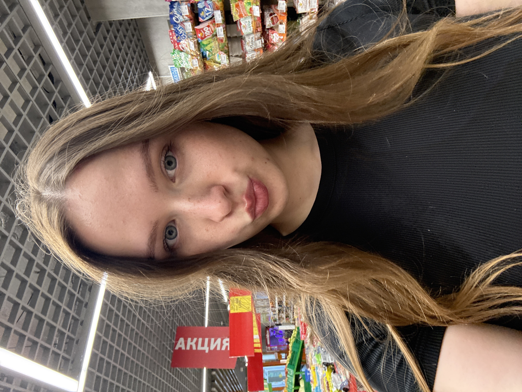 A young woman is featured in a close-up selfie, taken inside a supermarket located in Chaikovsky, Russia. She has long, light brown hair styled with a center part, light blue eyes, and fair skin displaying visible freckles across her forehead and cheeks. Her lips are tightly pursed in a "kissy face" expression. She is wearing a black, ribbed, short-sleeved top. The background reveals typical grocery store elements: to the left, several produce displays with various fruits and vegetables; to the right, tall shelving units stocked with numerous packaged snack items, including bags with Russian labels like "СУХАРИКИ" (crackers), "Перец" (pepper), and "Карри" (curry), along with price tags. Red promotional banners, one prominently displaying "АКЦИЯ" (PROMOTION) in Cyrillic, hang from the ceiling on the left. A partial blue sign on the right appears to show "ВЫХОД" (EXIT). The ceiling is a suspended grid pattern illuminated by long, rectangular fluorescent light fixtures.
