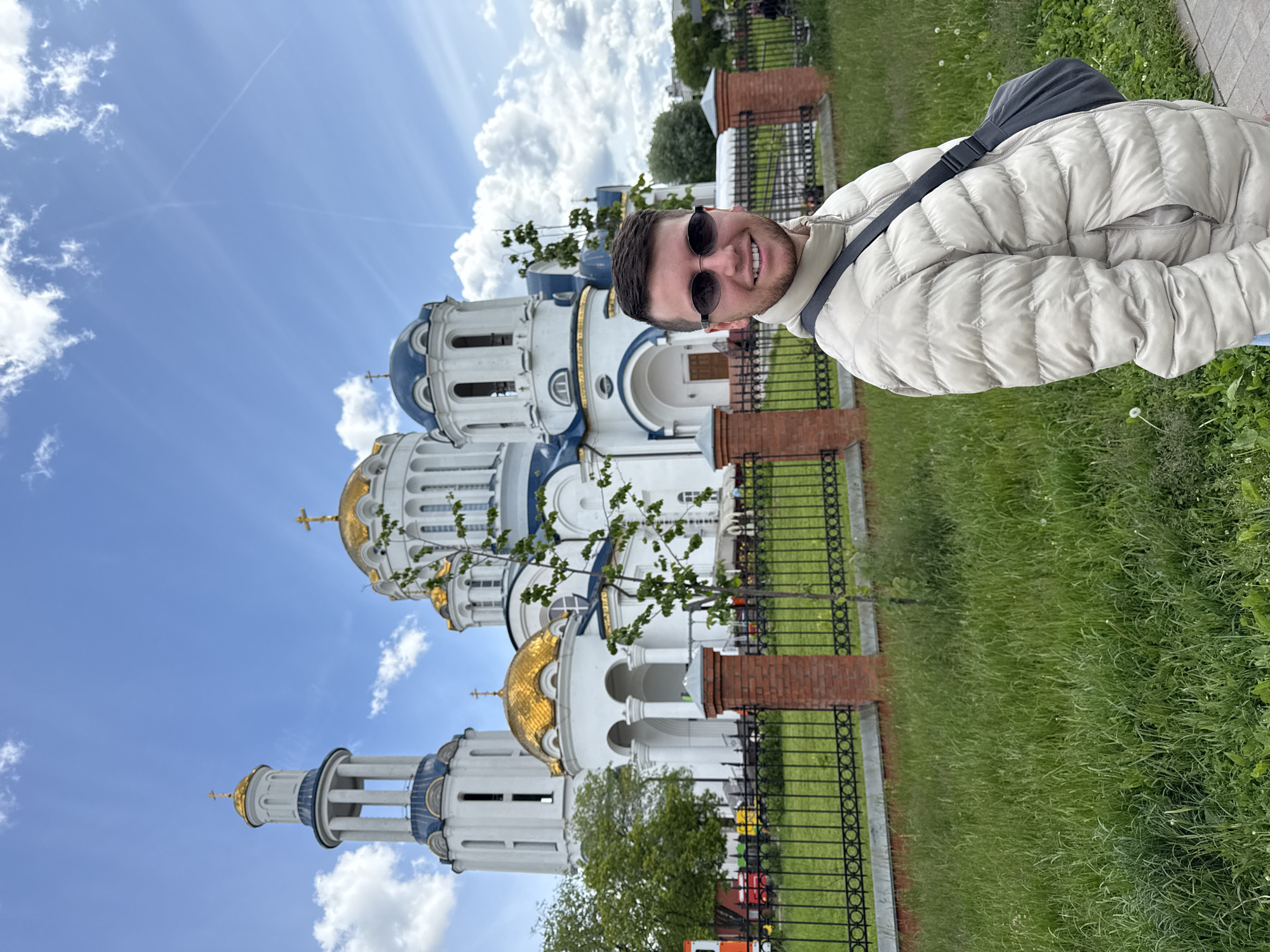 A man wearing a white puffer jacket and sunglasses stands in front of a Russian Orthodox church with blue and gold domes. The church is surrounded by a wrought-iron fence with brick pillars. The sky is blue with puffy white clouds. Green grass is visible in the foreground. The location is Moscow, Russia.