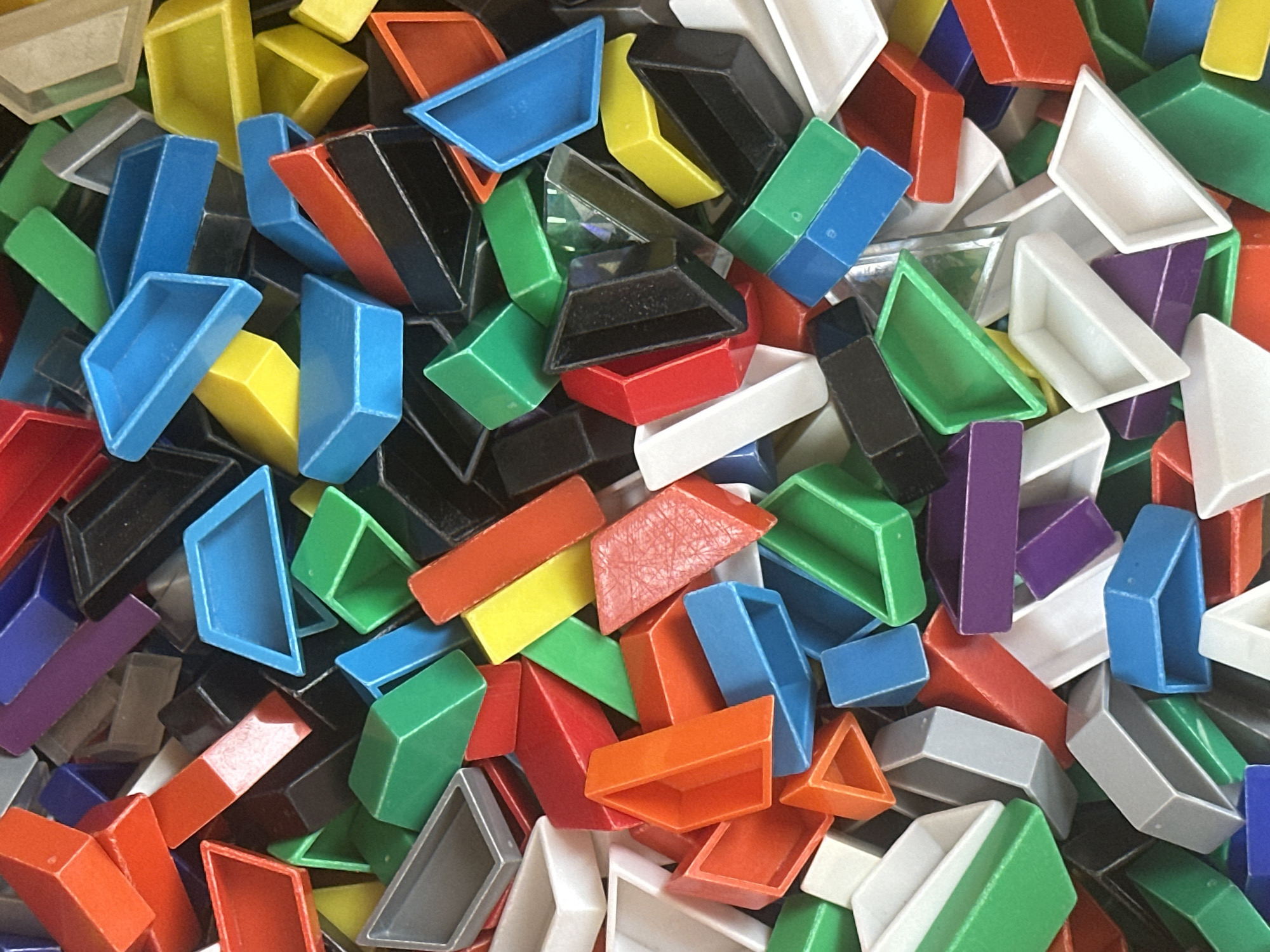 An overhead, close-up, full shot shows a pile of multi-colored geometric plastic toys. The toys are a mixture of trapezoids, triangles, and rectangular prisms, in red, blue, green, yellow, purple, black, and white. The objects are jumbled together, creating a chaotic, textured surface. The lighting is bright and even, casting no shadows. The background is entirely filled with the pile of toys.