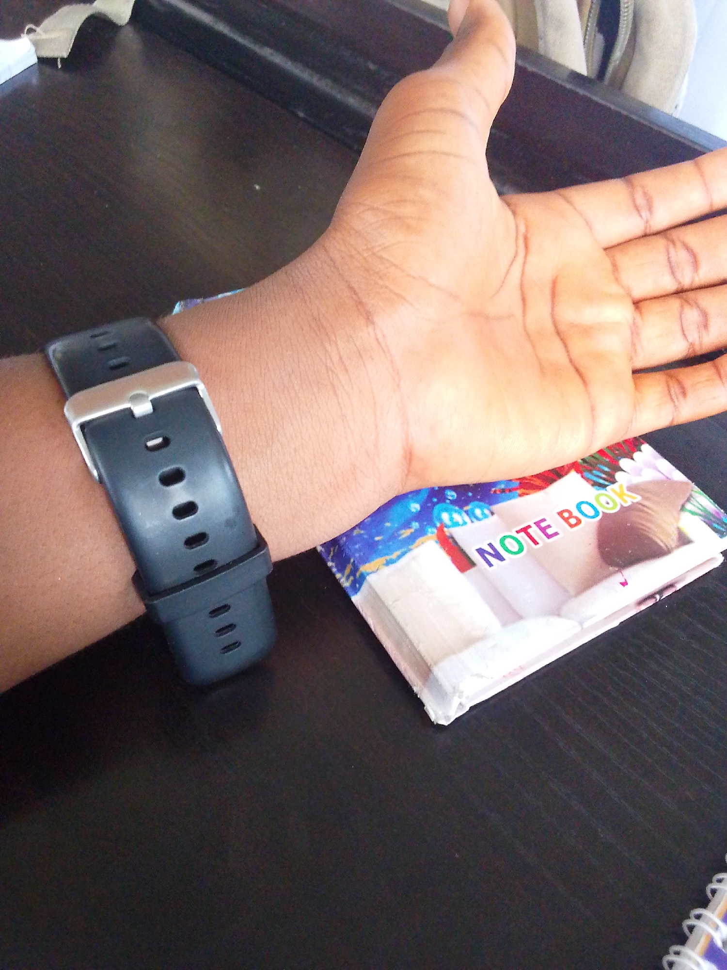 A person's left forearm and hand are visible, resting on a dark, textured surface, likely a desk. A black digital watch with a silver buckle is fastened around the wrist. In front of the forearm lies a small, colorful notebook with the words "NOTE BOOK" printed on its cover. The background shows a blurred object, possibly a bag or clothing, with a zipper. The scene is brightly lit, casting minimal shadows.
