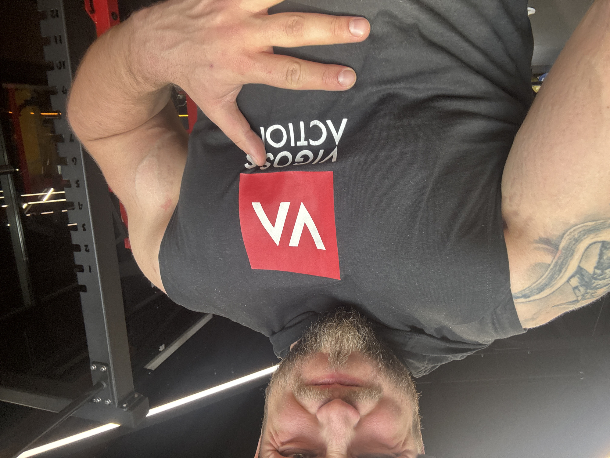 A man with a beard and a tattoo on his left arm is wearing a sleeveless black hoodie with a red and white logo. The logo features a red square with "VA" in white capital letters, and below it, "VIGOSS ACTION" in white capital letters. He is in a gym, with a power rack visible in the background. He is making a hand gesture with his right hand, with two fingers extended.