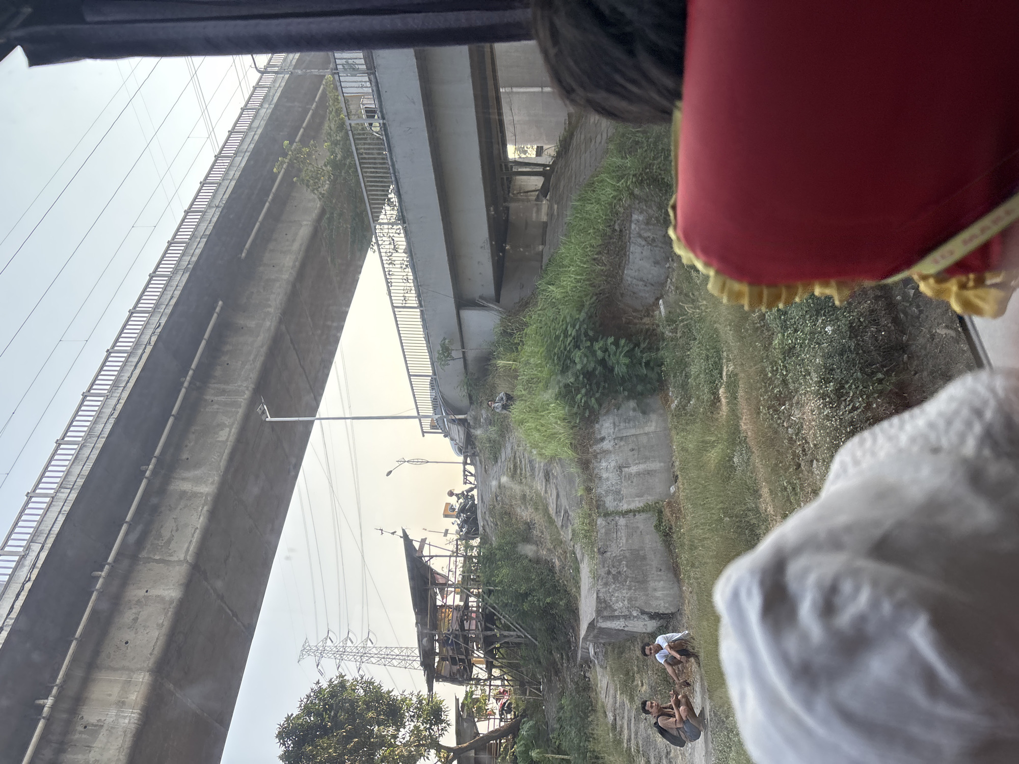 The image depicts an urban landscape in Jayamukti, Indonesia, characterized by an elevated highway structure and its immediate surroundings. In the foreground, a person's head and shoulder, covered by a white cloth, are visible, obscuring a portion of the scene. Adjacent to this, a red cushion with yellow trim is also in the foreground.
The midground reveals two young men squatting on a grassy incline. One is wearing shorts and a backpack, while the other is in shorts and a white t-shirt. Further up the incline, another man is seated on the grass. A dirt path winds its way up the slope. To the left, a makeshift structure made of wood and corrugated metal stands on stilts, with several motorcycles parked nearby. A tall electrical tower is visible in the distance.
The background is dominated by a multi-level concrete overpass or bridge system. The sky is clear and bright, suggesting it is daytime. The environment appears to be an area undergoing development or experiencing a blend of infrastructure and informal settlements.