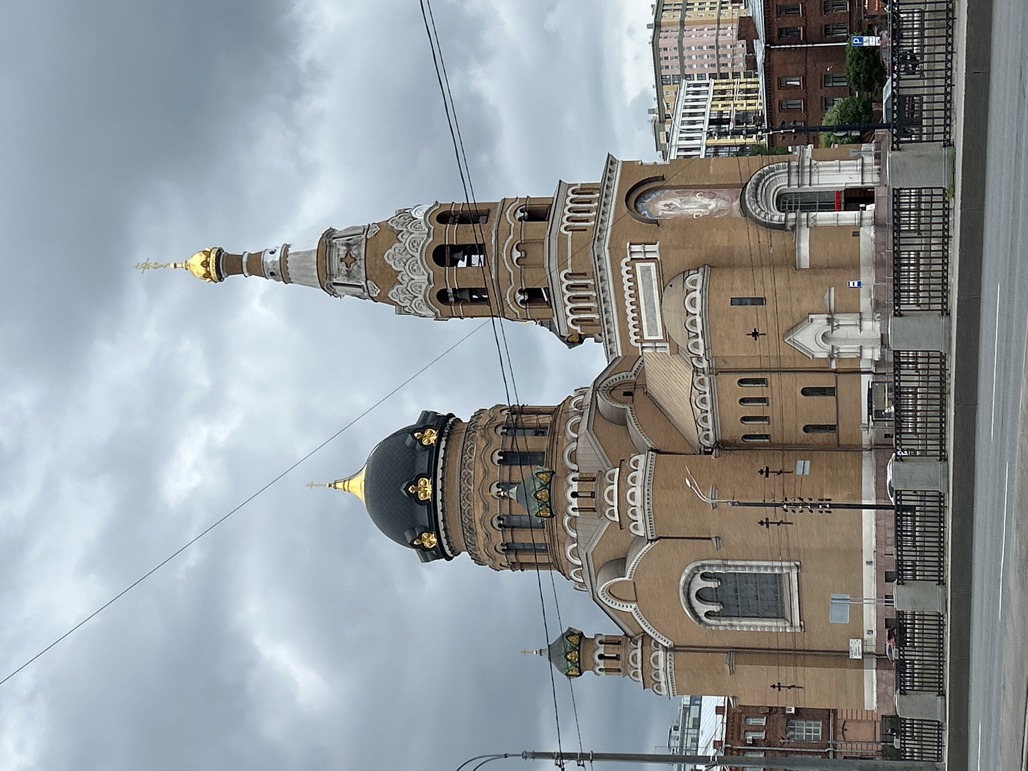 This is a vertically oriented, eye-level, medium shot of an ornate church building under a cloudy sky. The church has a dominant light brown facade, with multiple domes and decorative elements. The largest dome is dark, possibly black or dark green, with gold accents. To its left, a tall, slender tower with a golden spire pierces the sky. The tower has multiple levels with arched openings that appear to house bells. Smaller domes and spires are visible further down the building. The architecture features intricate detailing, including decorative moldings and arched windows. Religious imagery, possibly a fresco, is visible on the upper part of the building.
In the foreground, there is a paved road and a black metal fence separating the church from the road. Several power lines crisscross the sky. In the background, other buildings, some appearing to be residential or commercial, are visible. The sky is overcast, suggesting a cloudy day, possibly with impending rain, and the lighting is diffused. The scene appears to be set in an urban environment. No people are visible in the frame. The image has a slightly desaturated quality, consistent with a cloudy day.